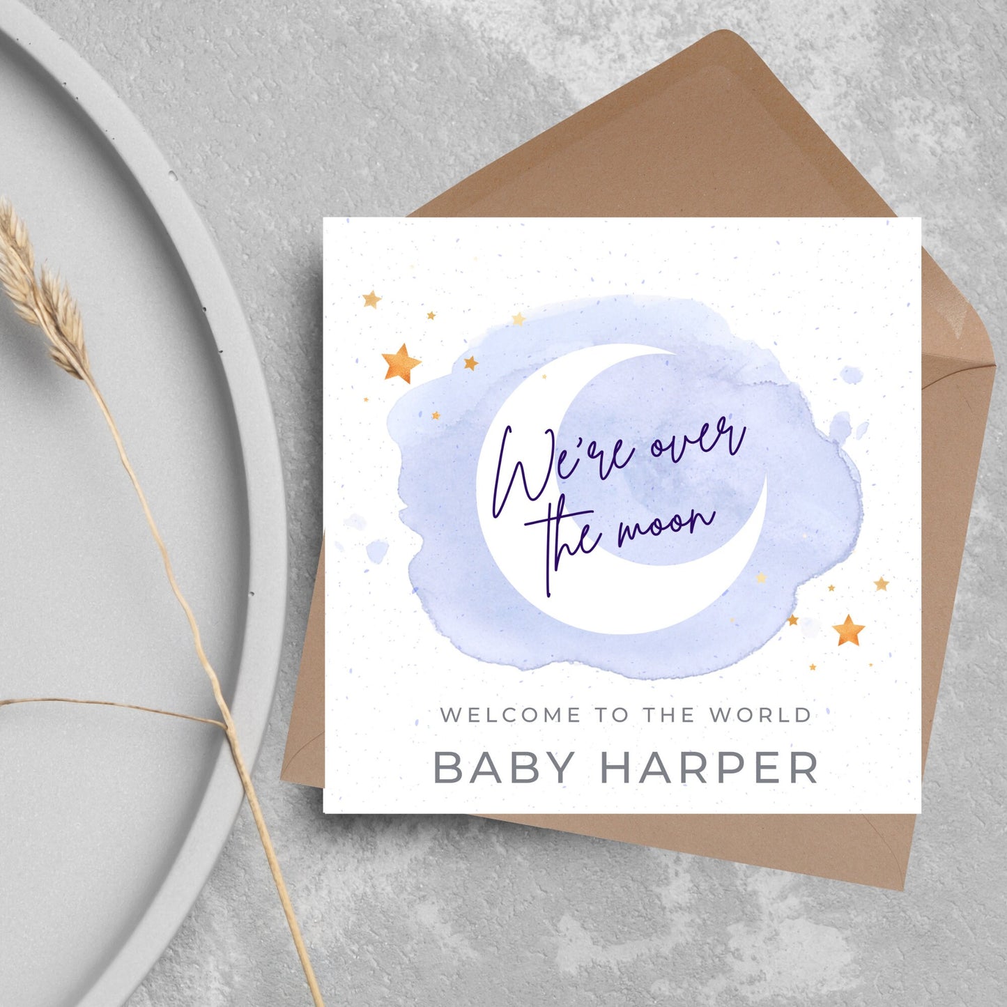 square card with a watercolour light blue background with a white crescent moon and yellow stars with navy blue hand script that reads - we're over the moon, and at the bottom in grey text welcome to the world baby harper