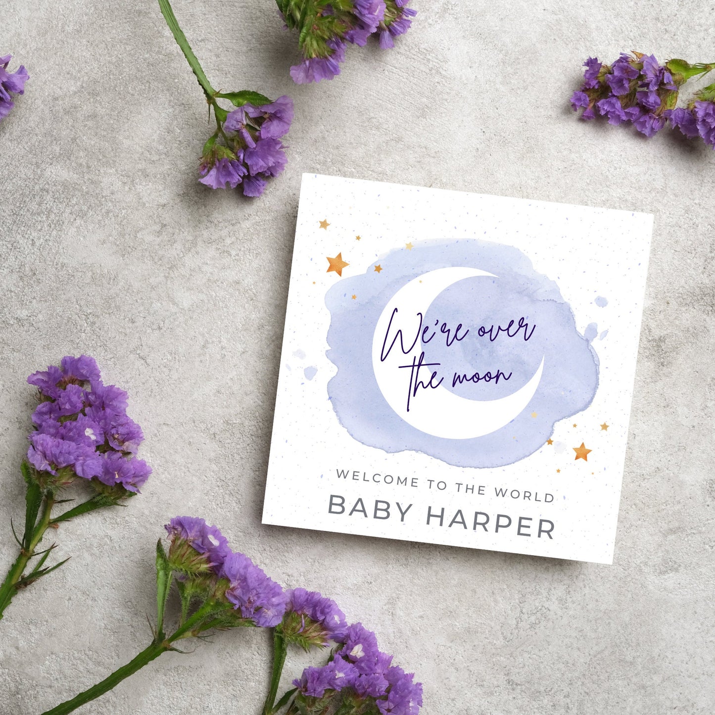 square card with a watercolour light blue background with a white crescent moon and yellow stars with navy blue hand script that reads - we're over the moon, and at the bottom in grey text welcome to the world baby harper
