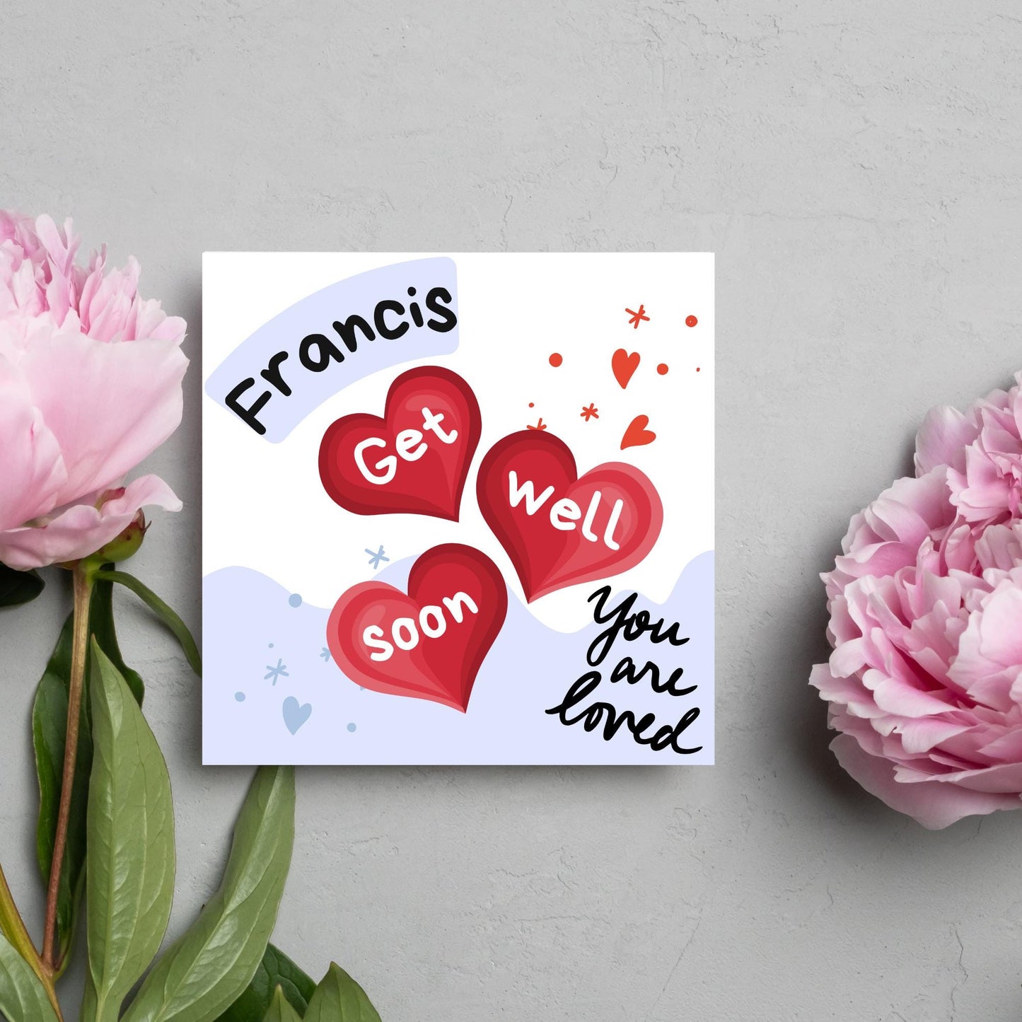 square card with white and blue background. 3 red hearts are in the centre of the card with get well soon overlayed in white, the top corner has curved text with the recipients name and in the bottom corner the text you are loved in black cursive