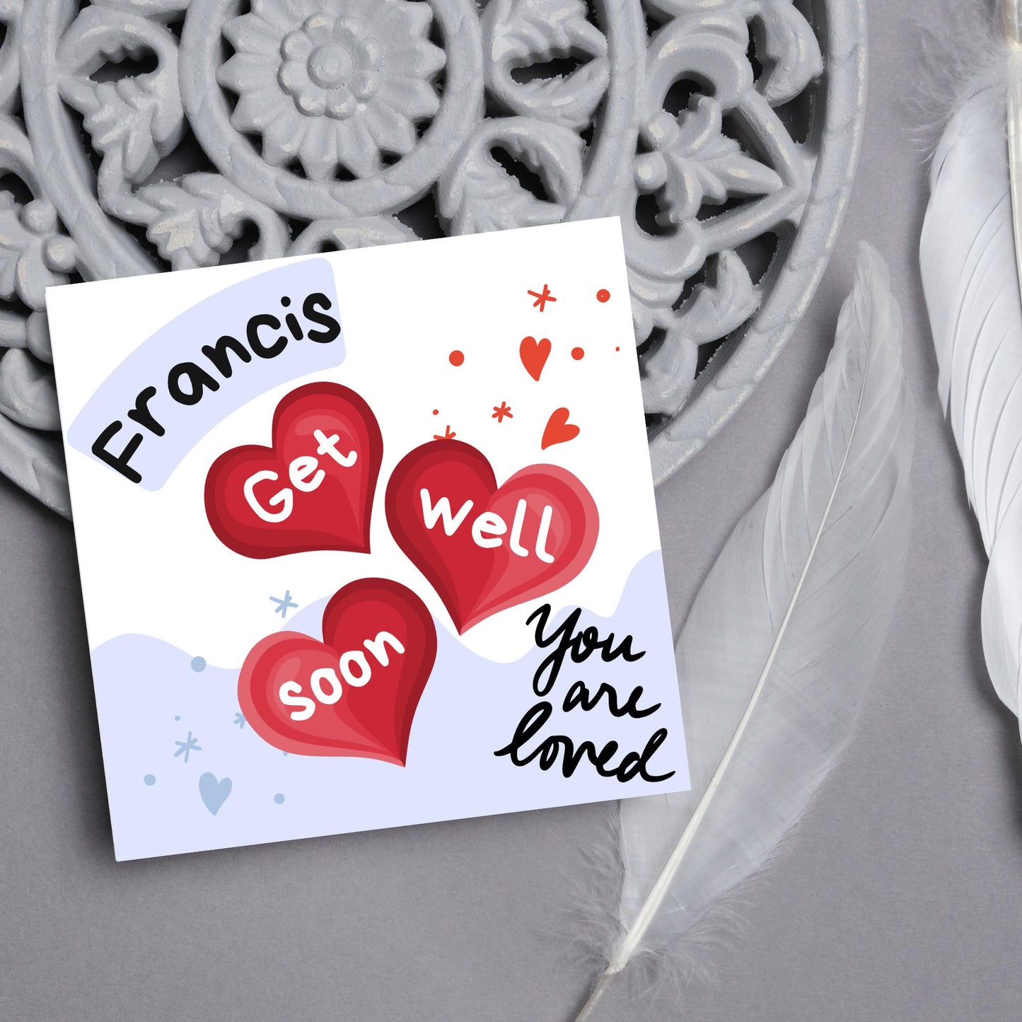 square card with white and blue background. 3 red hearts are in the centre of the card with get well soon overlayed in white, the top corner has curved text with the recipients name and in the bottom corner the text you are loved in black cursive