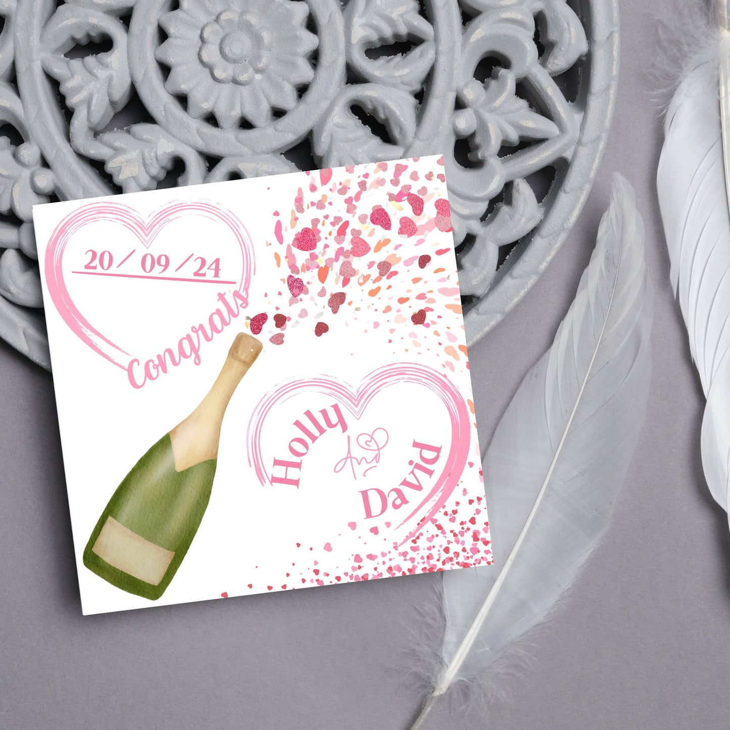 square wedding card with a bottle of champagne bursting with hearts in varying shades of pink. Above the bottle is a heart that holds the date and says contrats, and below is a repeated heart with the names of the couple inside