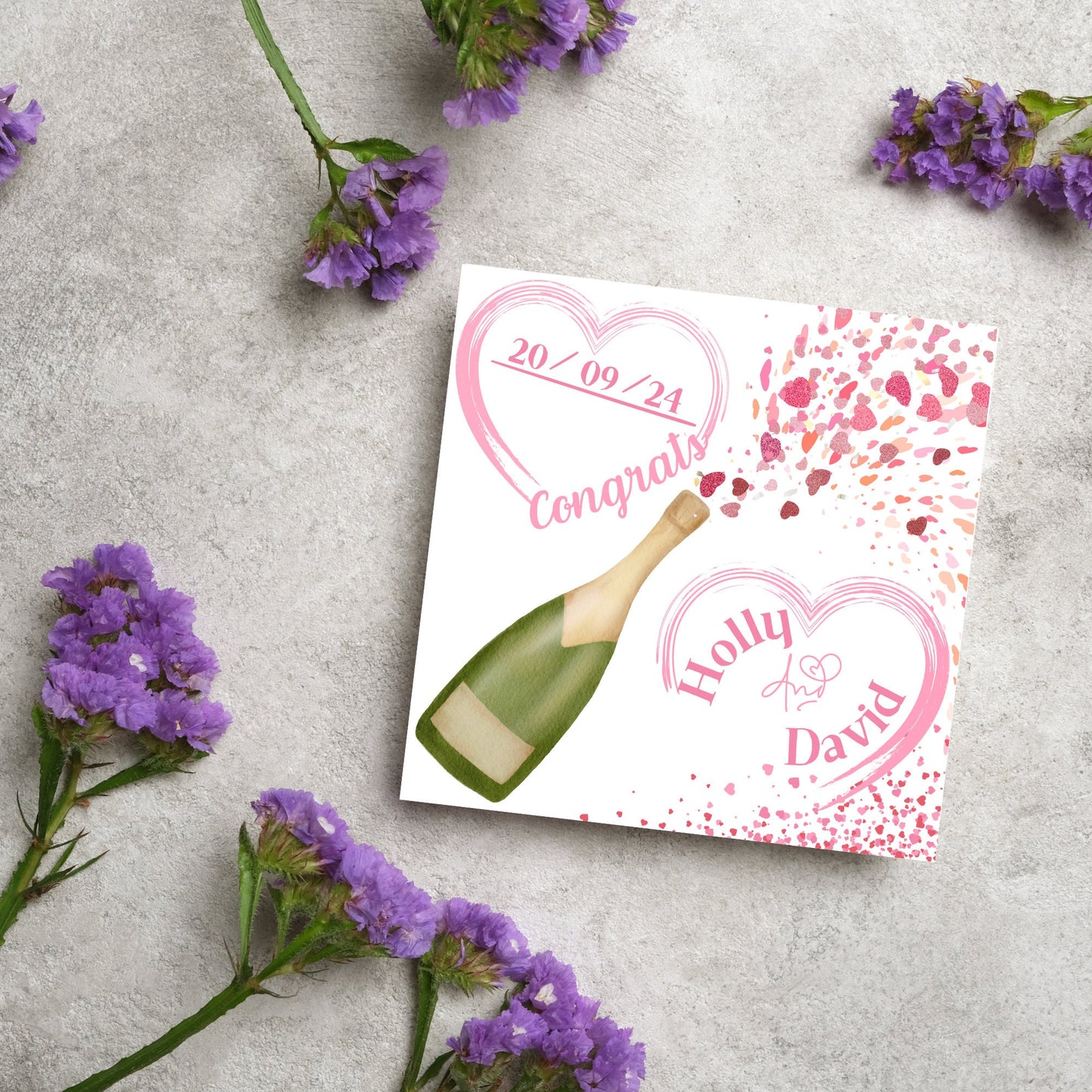 square wedding card with a bottle of champagne bursting with hearts in varying shades of pink. Above the bottle is a heart that holds the date and says contrats, and below is a repeated heart with the names of the couple inside