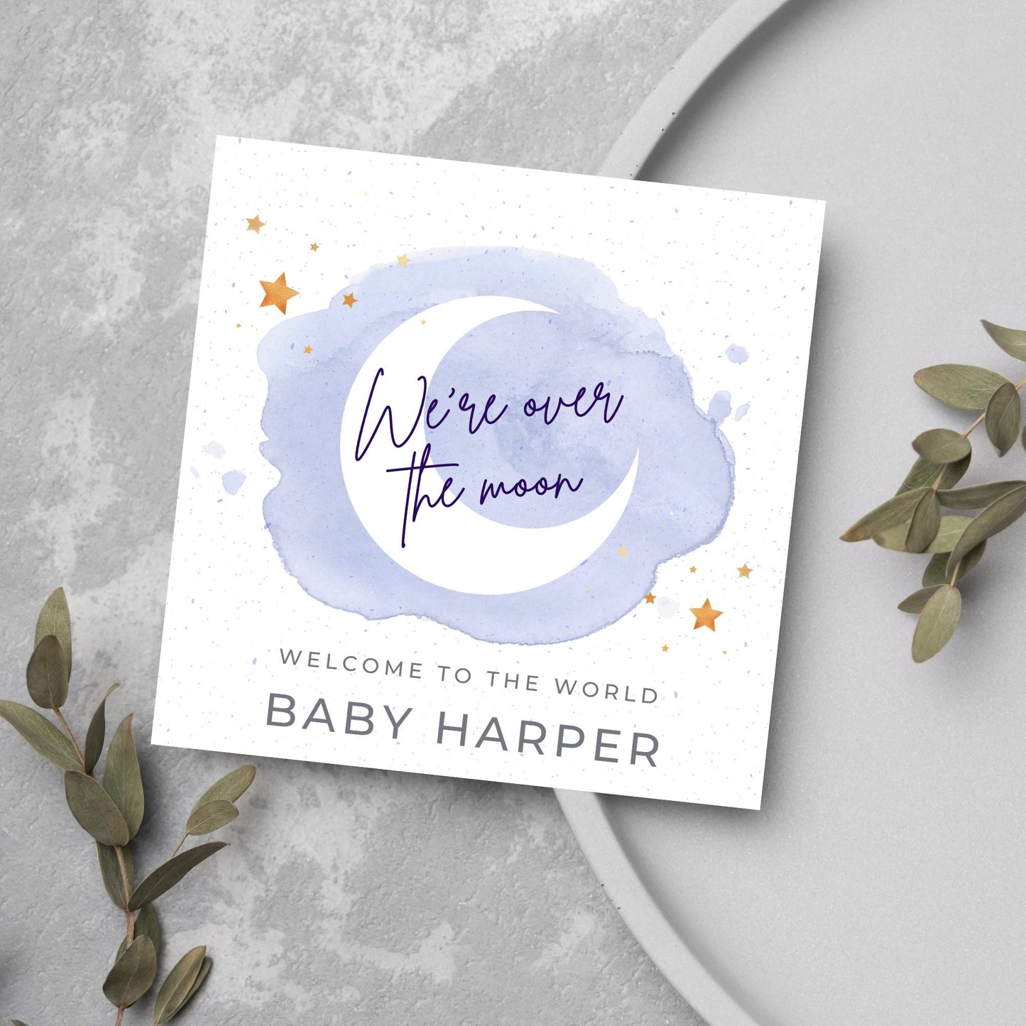 square card with a watercolour light blue background with a white crescent moon and yellow stars with navy blue hand script that reads - we're over the moon, and at the bottom in grey text welcome to the world baby harper