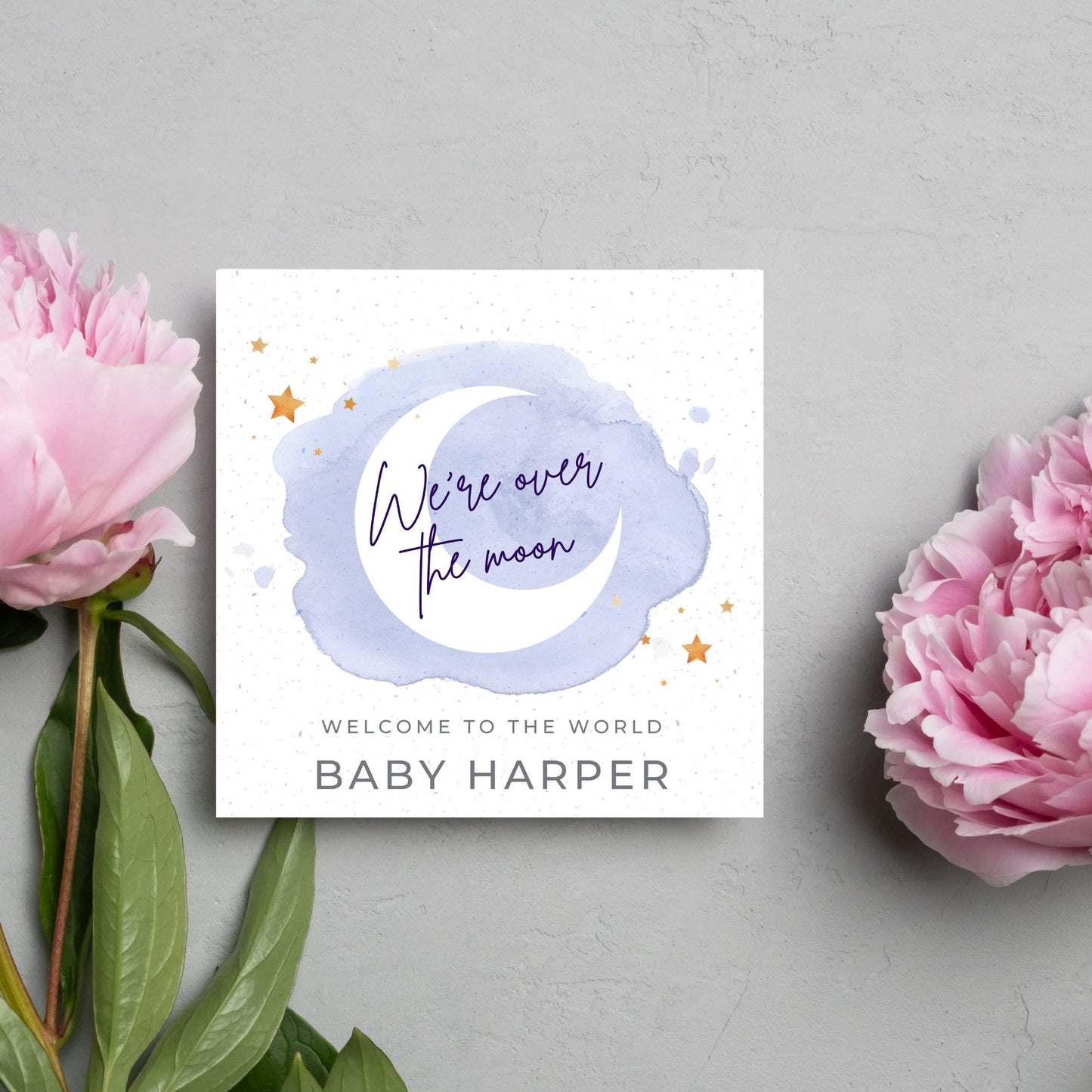 square card with a watercolour light blue background with a white crescent moon and yellow stars with navy blue hand script that reads - we're over the moon, and at the bottom in grey text welcome to the world baby harper