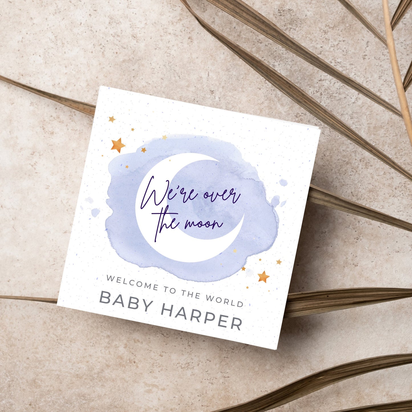 square card with a watercolour light blue background with a white crescent moon and yellow stars with navy blue hand script that reads - we're over the moon, and at the bottom in grey text welcome to the world baby harper
