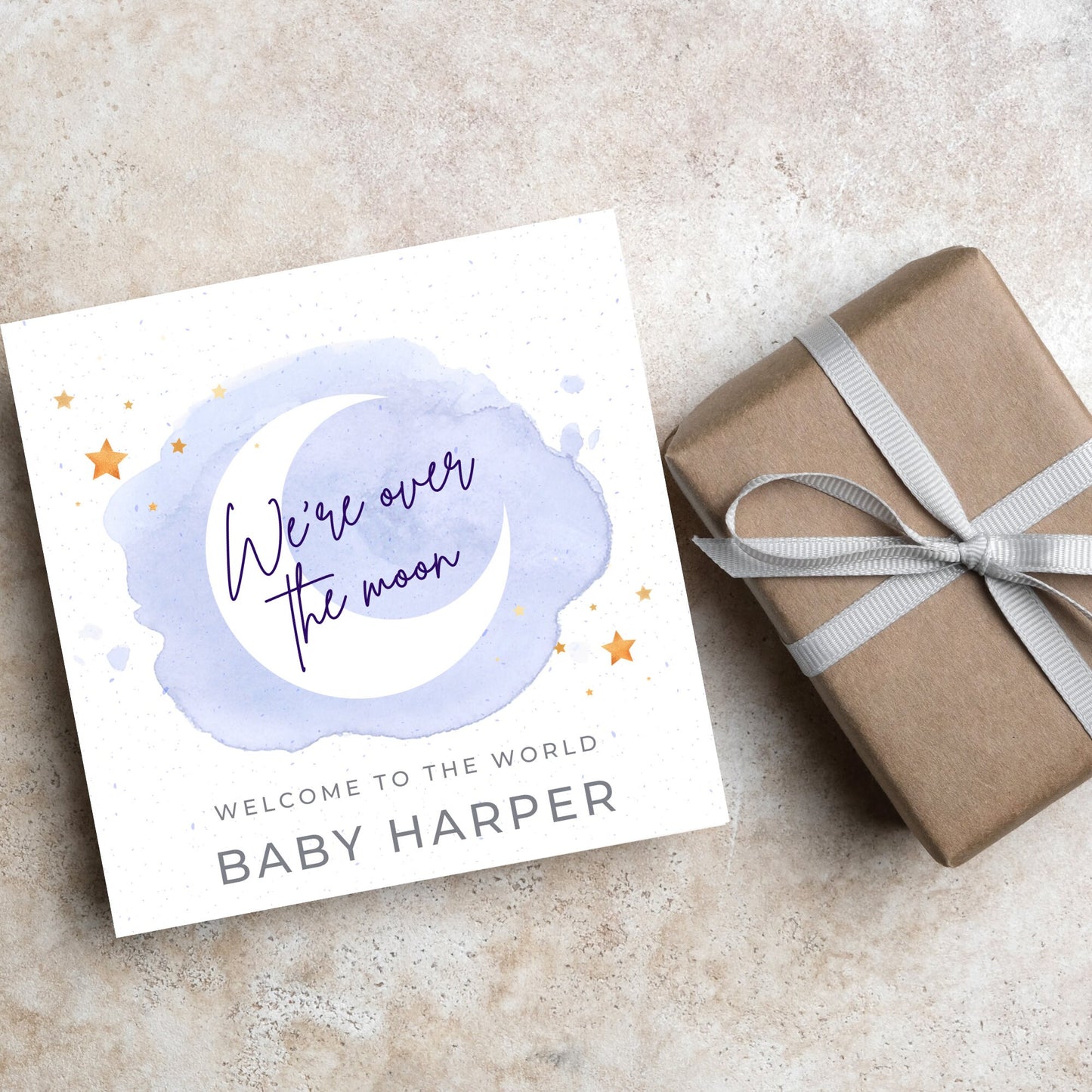 square card with a watercolour light blue background with a white crescent moon and yellow stars with navy blue hand script that reads - we're over the moon, and at the bottom in grey text welcome to the world baby harper