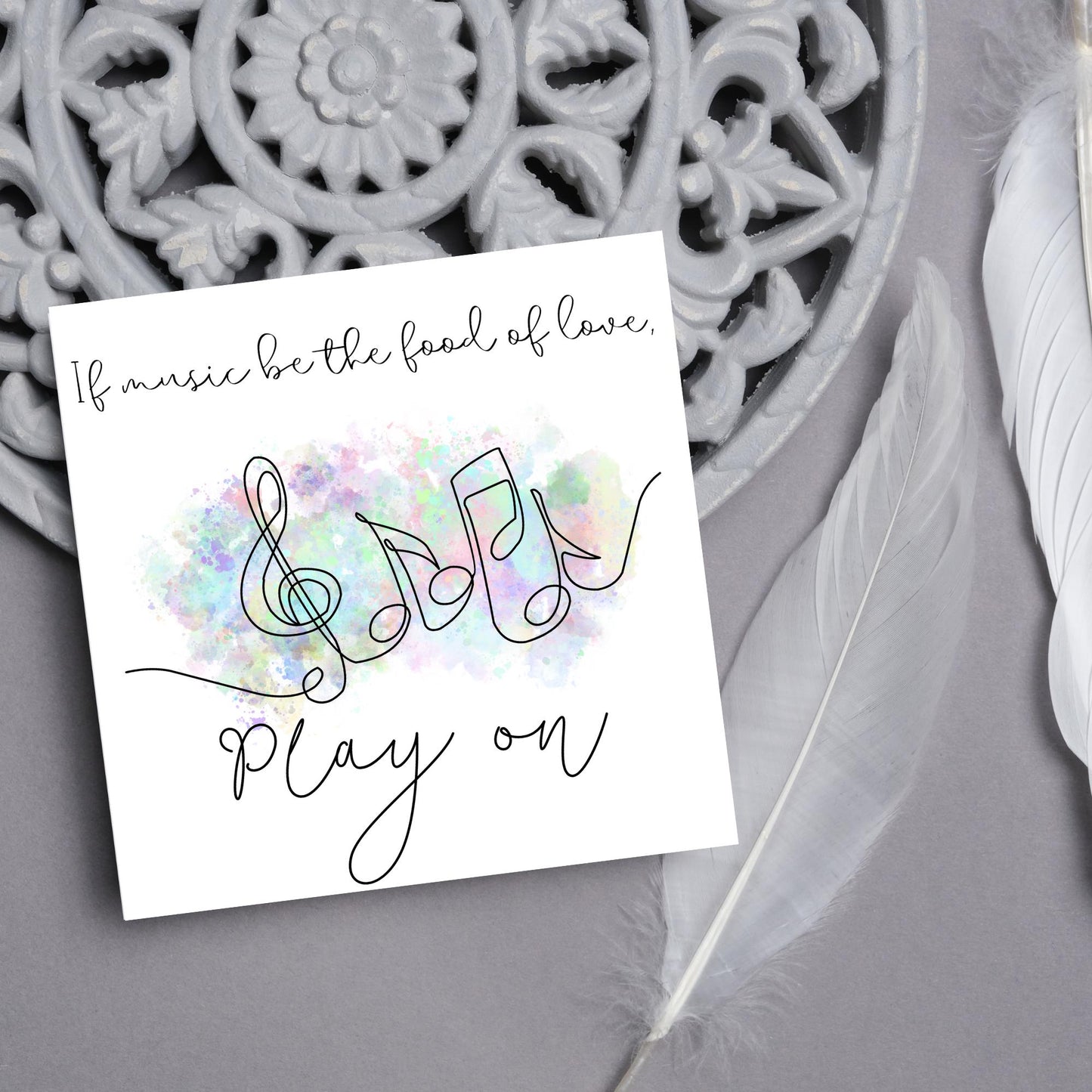 White square card with a one line drawing of musical notes that look like they are dancing in front of a pastel rainbow watercolour cloud. Cursive text reads, If music be the food of love, play on