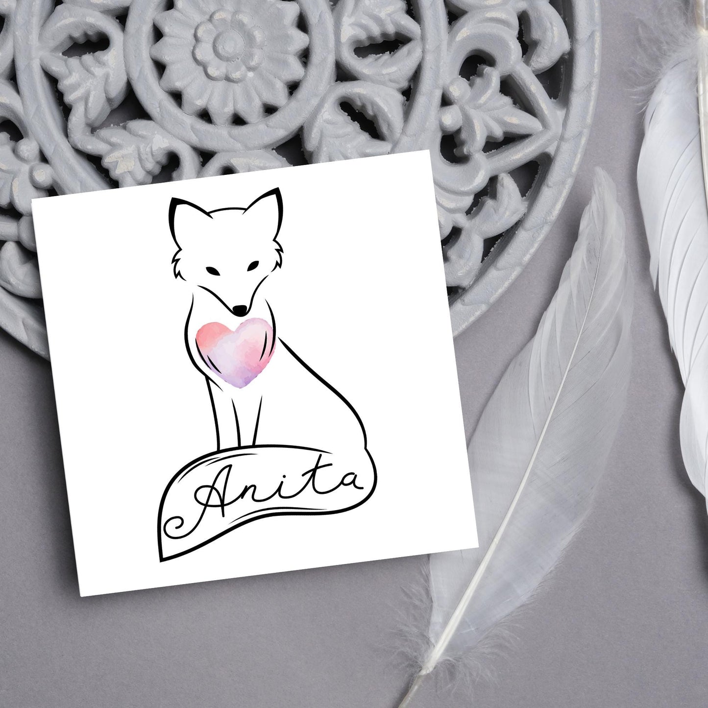 Square card with simple black line drawing of a fox sitting with its tail wrapped around its feet. It has a pink watercolour heart on its chest and personalised with a name on the tail in a calligraphy font