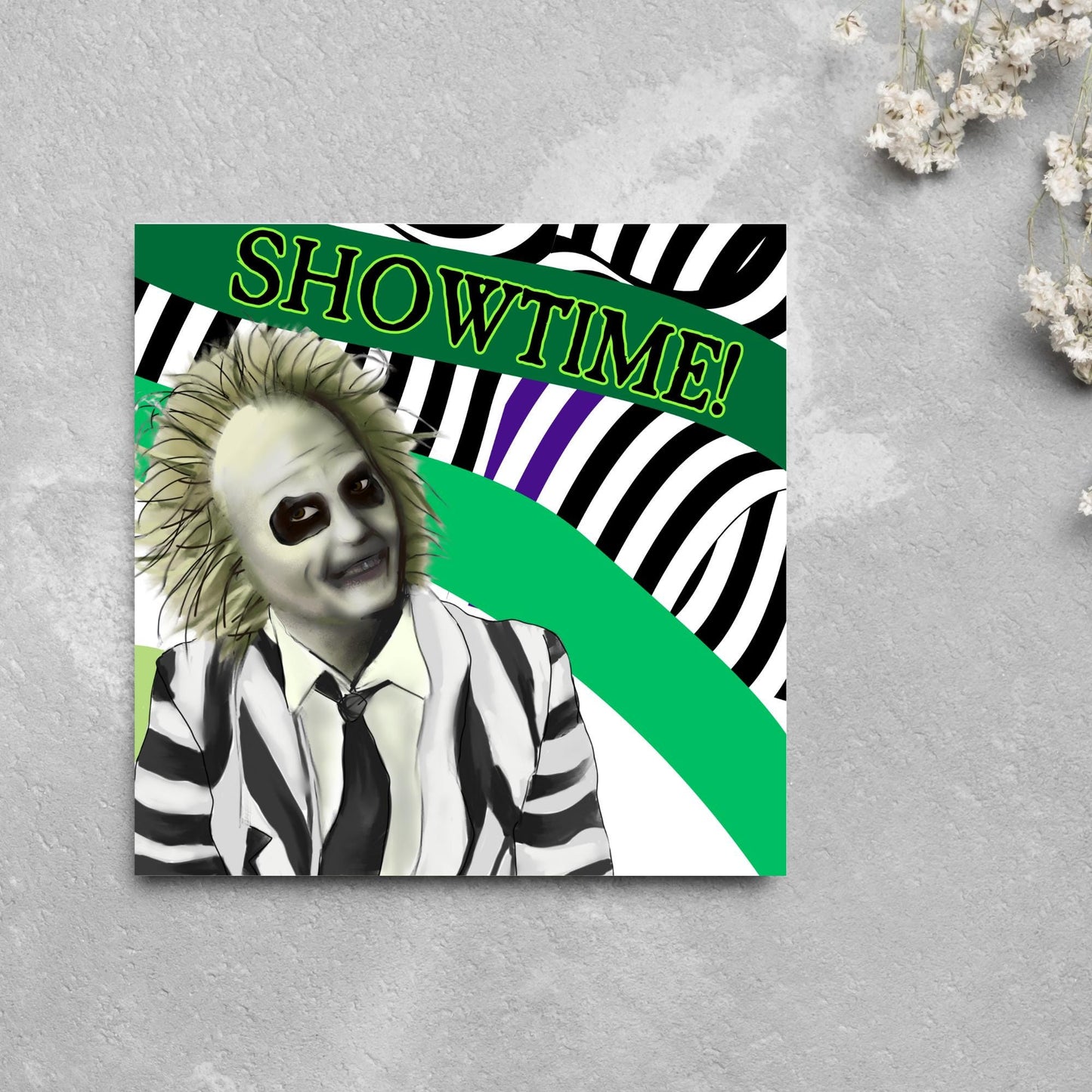 square greeting card with a drawn image of beetlejuice grinning in from of a black, green and purple striped background. On one of the biggest stripes is the word showtime in bold capital letters in a font similar to the movie title