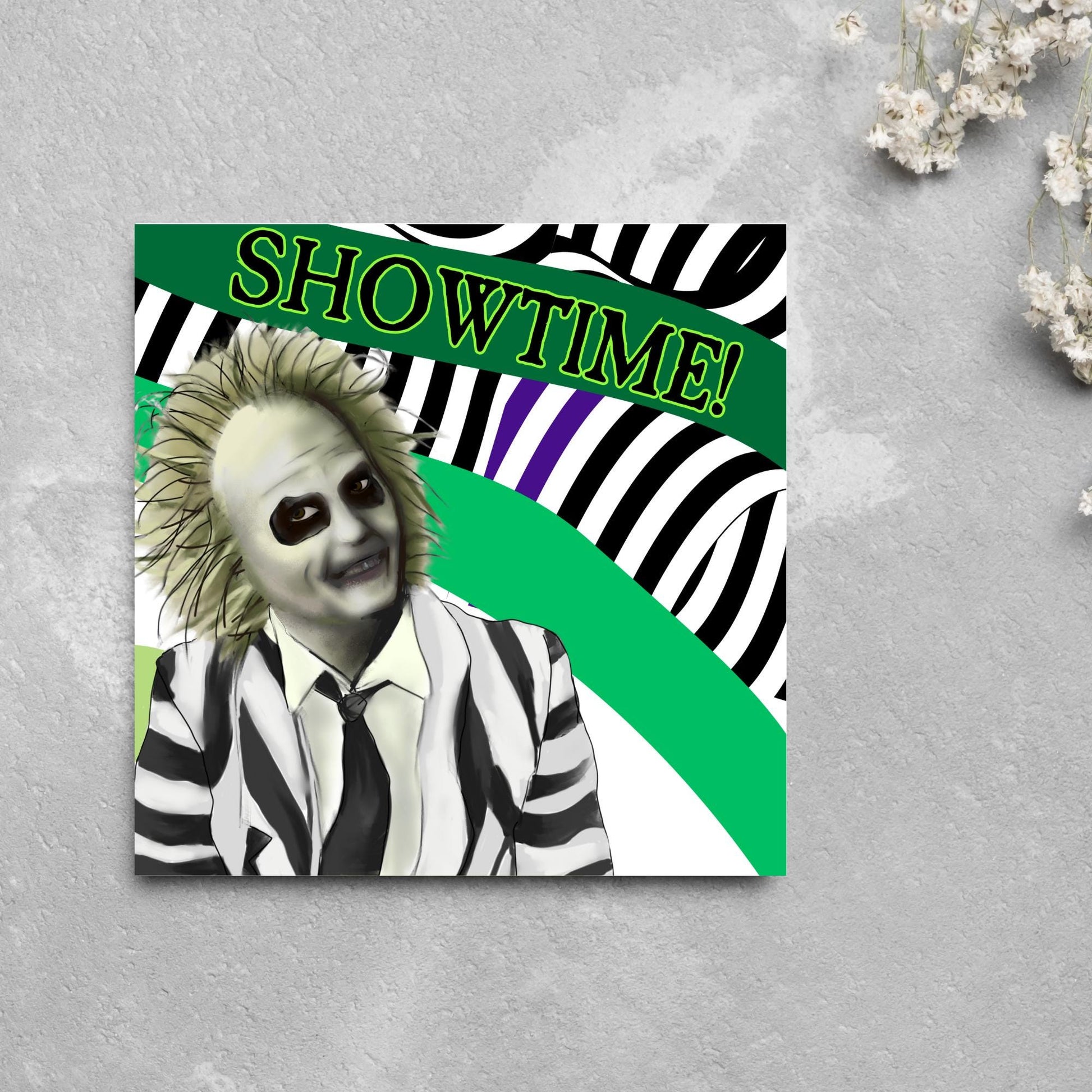 square greeting card with a drawn image of beetlejuice grinning in from of a black, green and purple striped background. On one of the biggest stripes is the word showtime in bold capital letters in a font similar to the movie title