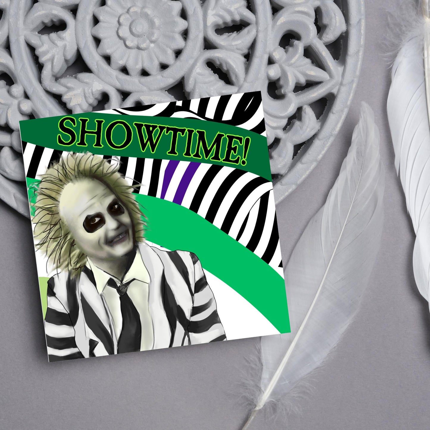 square greeting card with a drawn image of beetlejuice grinning in from of a black, green and purple striped background. On one of the biggest stripes is the word showtime in bold capital letters in a font similar to the movie title
