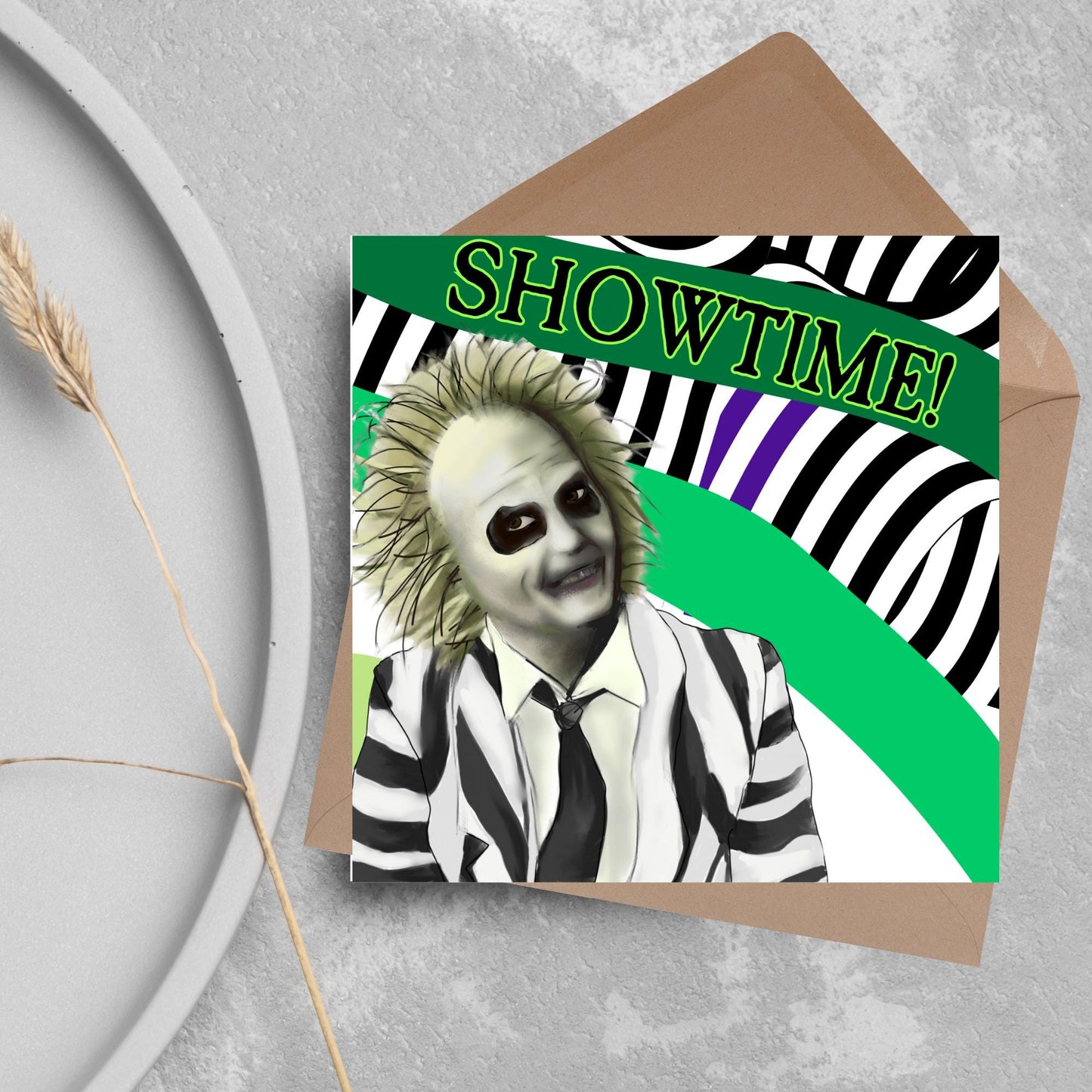 square greeting card with a drawn image of beetlejuice grinning in from of a black, green and purple striped background. On one of the biggest stripes is the word showtime in bold capital letters in a font similar to the movie title