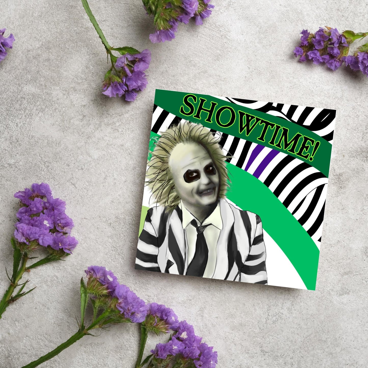 square greeting card with a drawn image of beetlejuice grinning in from of a black, green and purple striped background. On one of the biggest stripes is the word showtime in bold capital letters in a font similar to the movie title