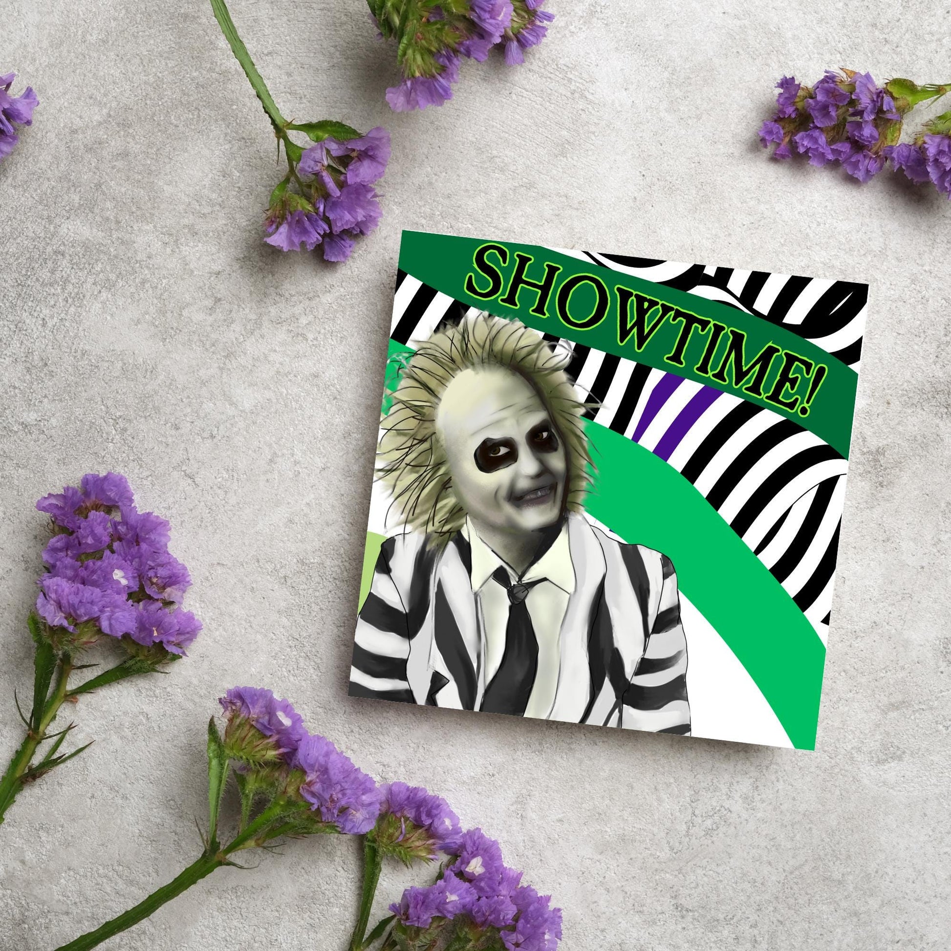 square greeting card with a drawn image of beetlejuice grinning in from of a black, green and purple striped background. On one of the biggest stripes is the word showtime in bold capital letters in a font similar to the movie title