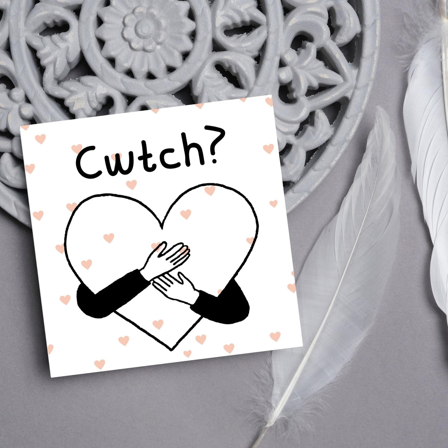 Square Greeting card small with subtle pink hearts in the background of a line drawn heart with hands hugging it and the word CWTCH? above it in handwritten style text