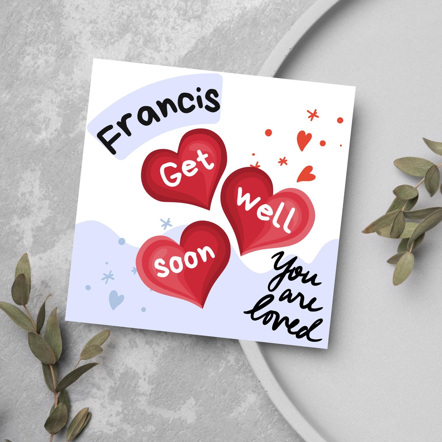 square card with white and blue background. 3 red hearts are in the centre of the card with get well soon overlayed in white, the top corner has curved text with the recipients name and in the bottom corner the text you are loved in black cursive