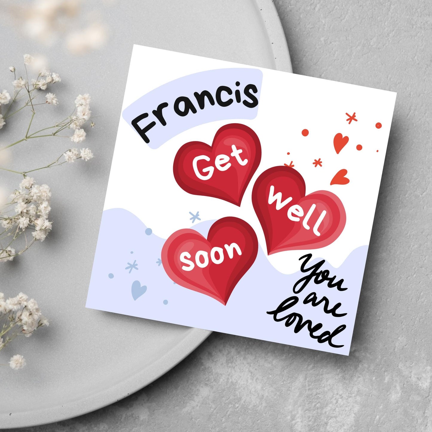 square card with white and blue background. 3 red hearts are in the centre of the card with get well soon overlayed in white, the top corner has curved text with the recipients name and in the bottom corner the text you are loved in black cursive