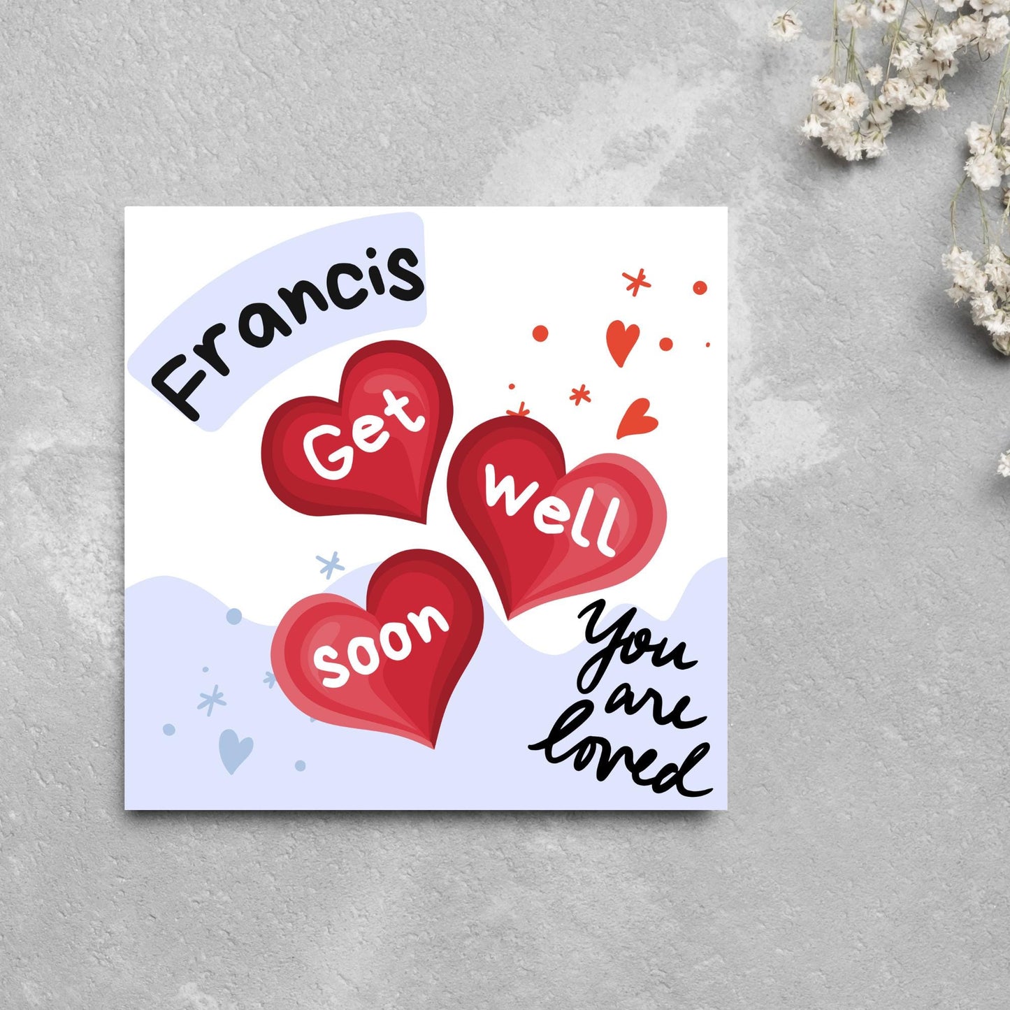 square card with white and blue background. 3 red hearts are in the centre of the card with get well soon overlayed in white, the top corner has curved text with the recipients name and in the bottom corner the text you are loved in black cursive