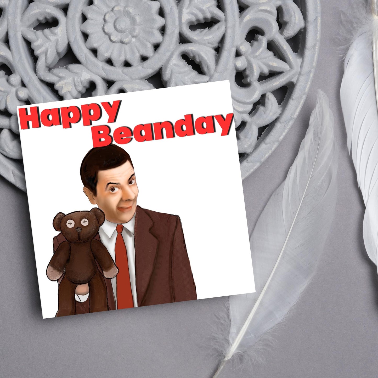 square card with a hand drawn image of Mr bean holding his teddy pooky with a cheeky smile on his face with the words Happy Beanday above in bold red text