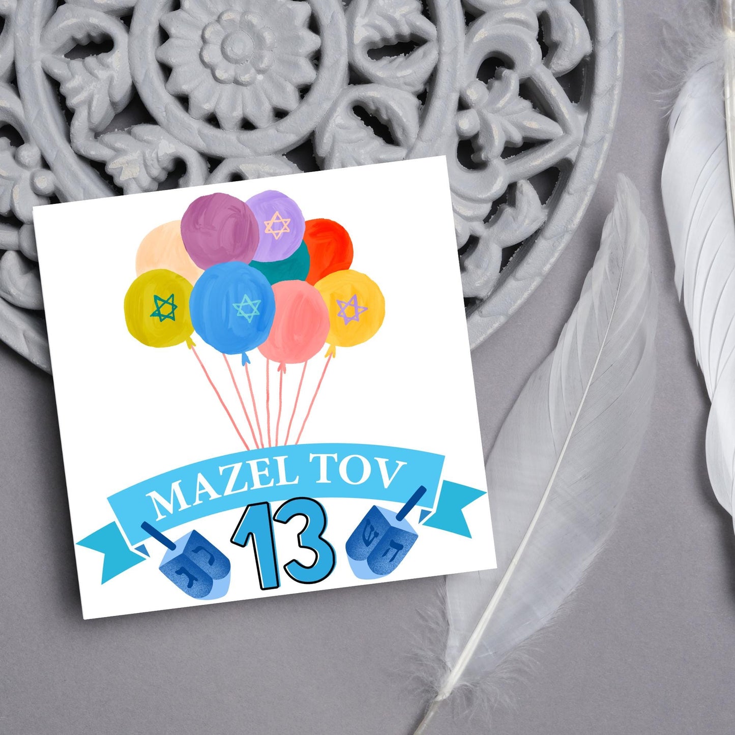 square greetings card with a bunch of watercolour design balloons decorated with stars of david. Below is a blue banner which reads mazel tov and the number 13 between a dreidel toy