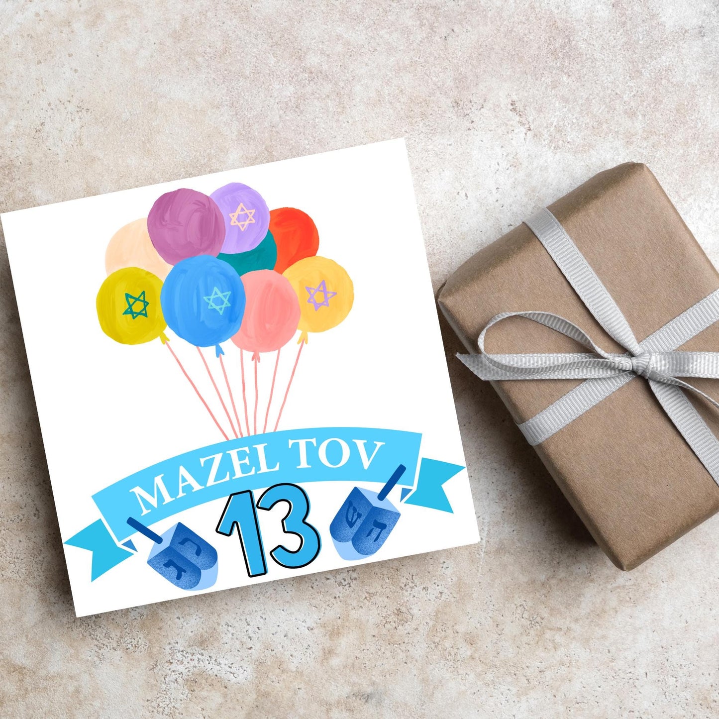 square greetings card with a bunch of watercolour design balloons decorated with stars of david. Below is a blue banner which reads mazel tov and the number 13 between a dreidel toy
