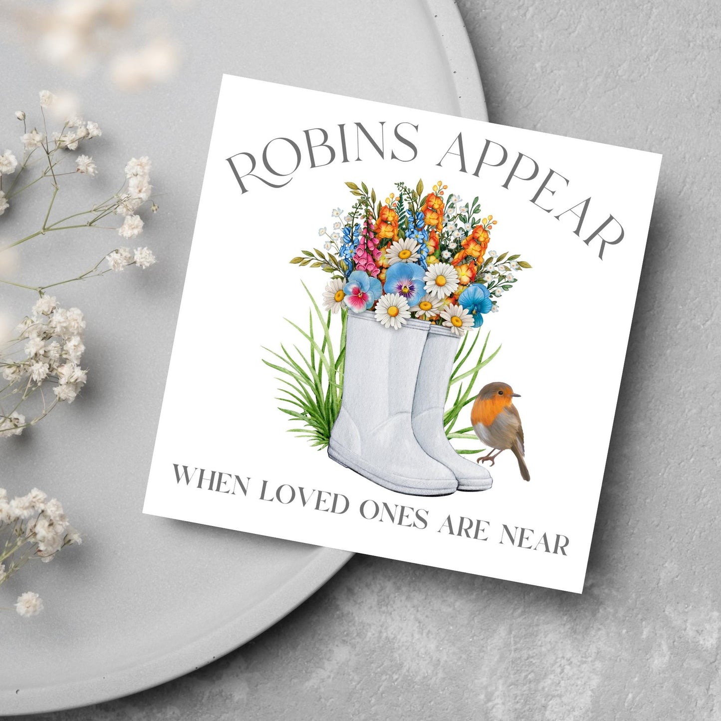 Square card with a watercolour image of a robin sitting on a pair of wellington boots that at filled with flowers. Elegant text reads Robins Appear when loved ones are near
