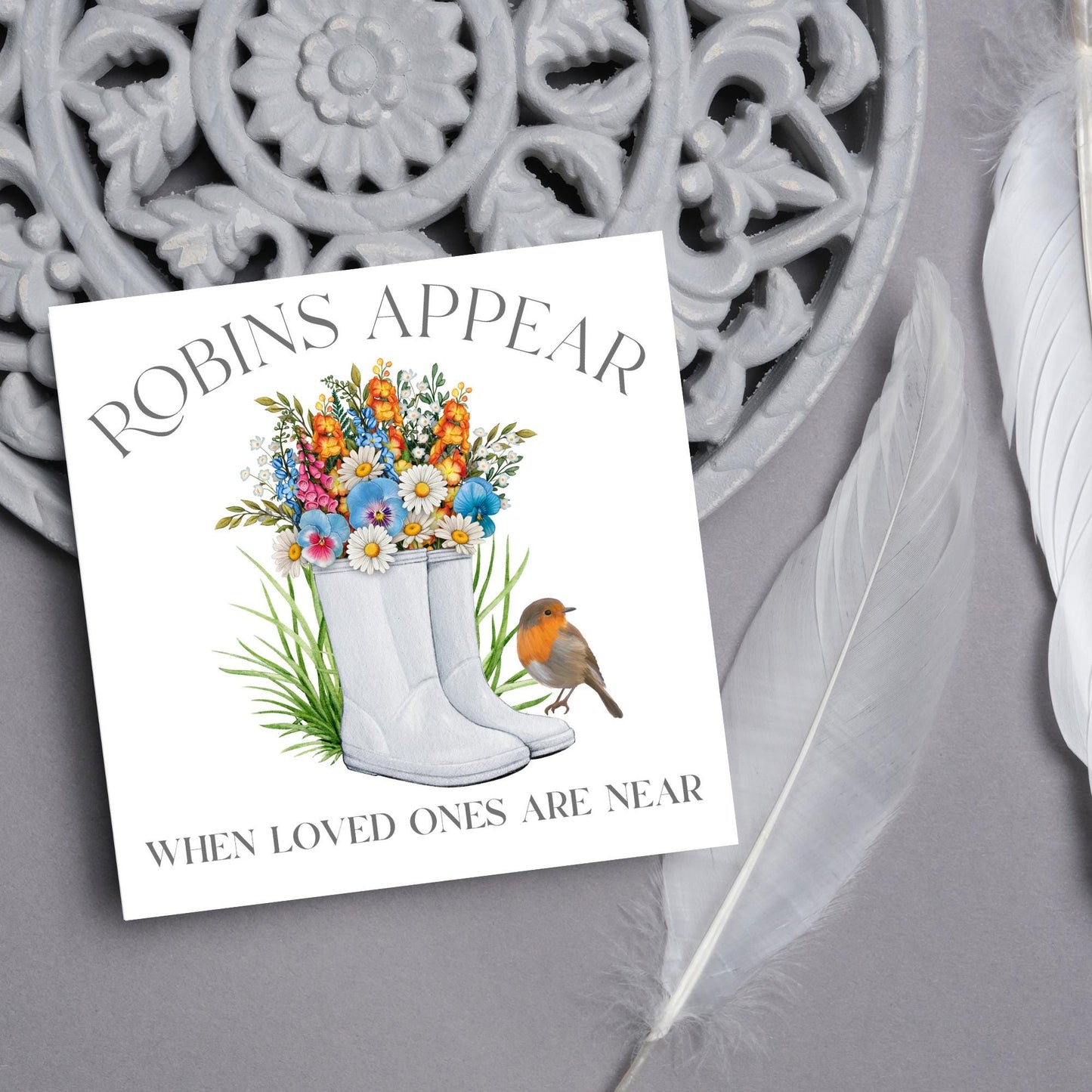 Square card with a watercolour image of a robin sitting on a pair of wellington boots that at filled with flowers. Elegant text reads Robins Appear when loved ones are near