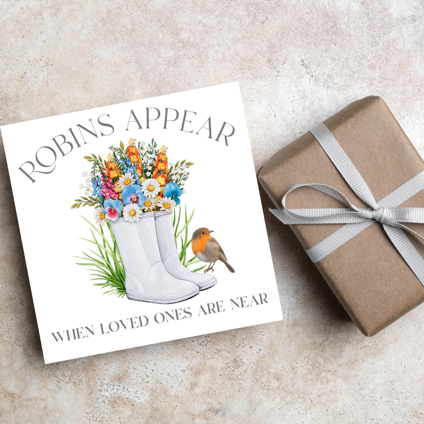 Square card with a watercolour image of a robin sitting on a pair of wellington boots that at filled with flowers. Elegant text reads Robins Appear when loved ones are near