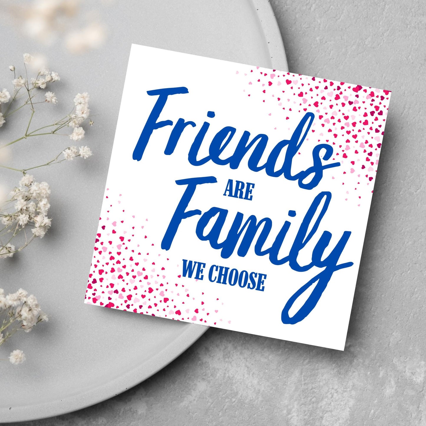 Square card with small hearts on opposite corners in shades of pink and blue writing in the centre that reads Friends are Family we choose