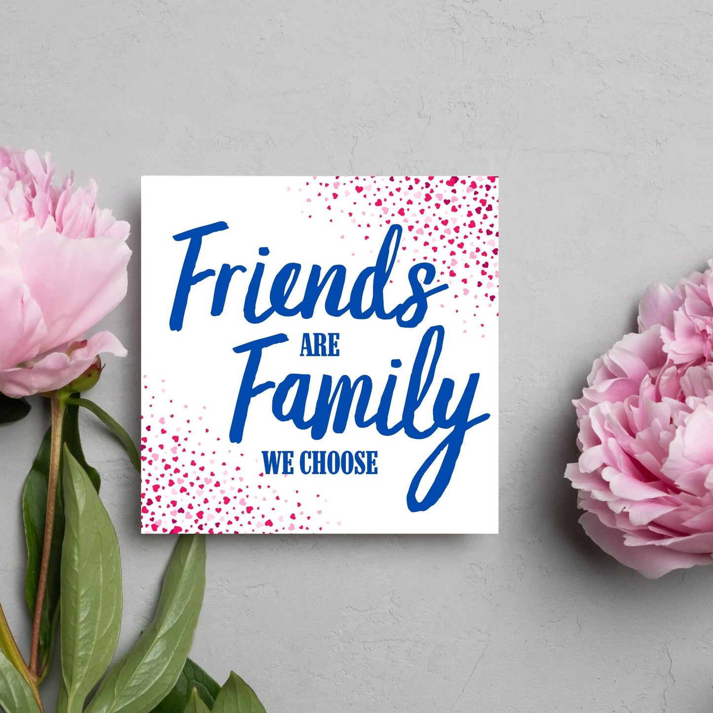 Square card with small hearts on opposite corners in shades of pink and blue writing in the centre that reads Friends are Family we choose