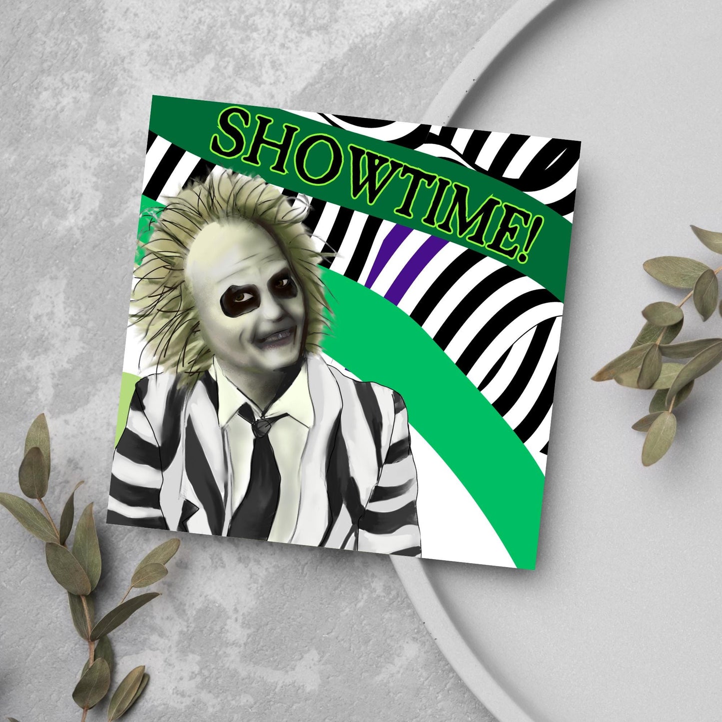 square greeting card with a drawn image of beetlejuice grinning in from of a black, green and purple striped background. On one of the biggest stripes is the word showtime in bold capital letters in a font similar to the movie title