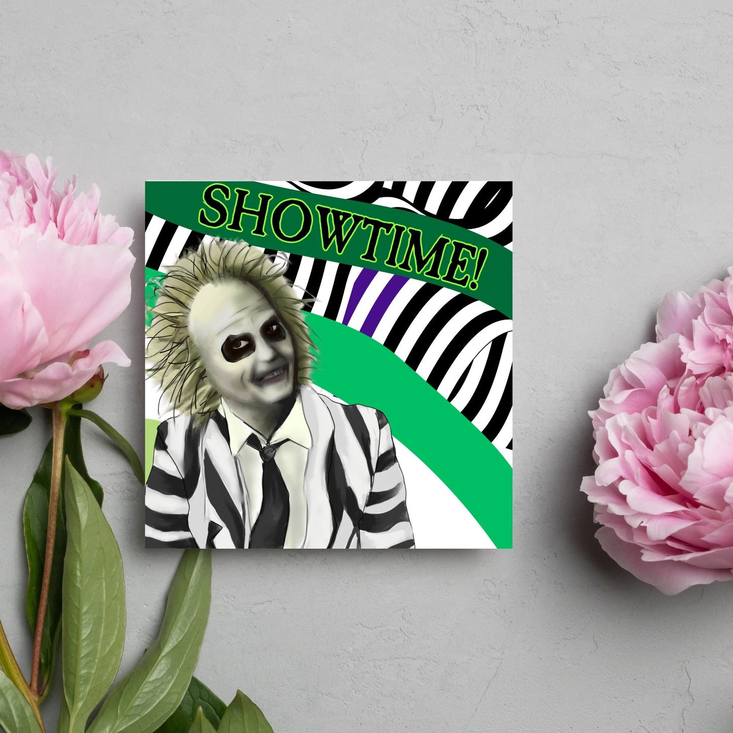 square greeting card with a drawn image of beetlejuice grinning in from of a black, green and purple striped background. On one of the biggest stripes is the word showtime in bold capital letters in a font similar to the movie title