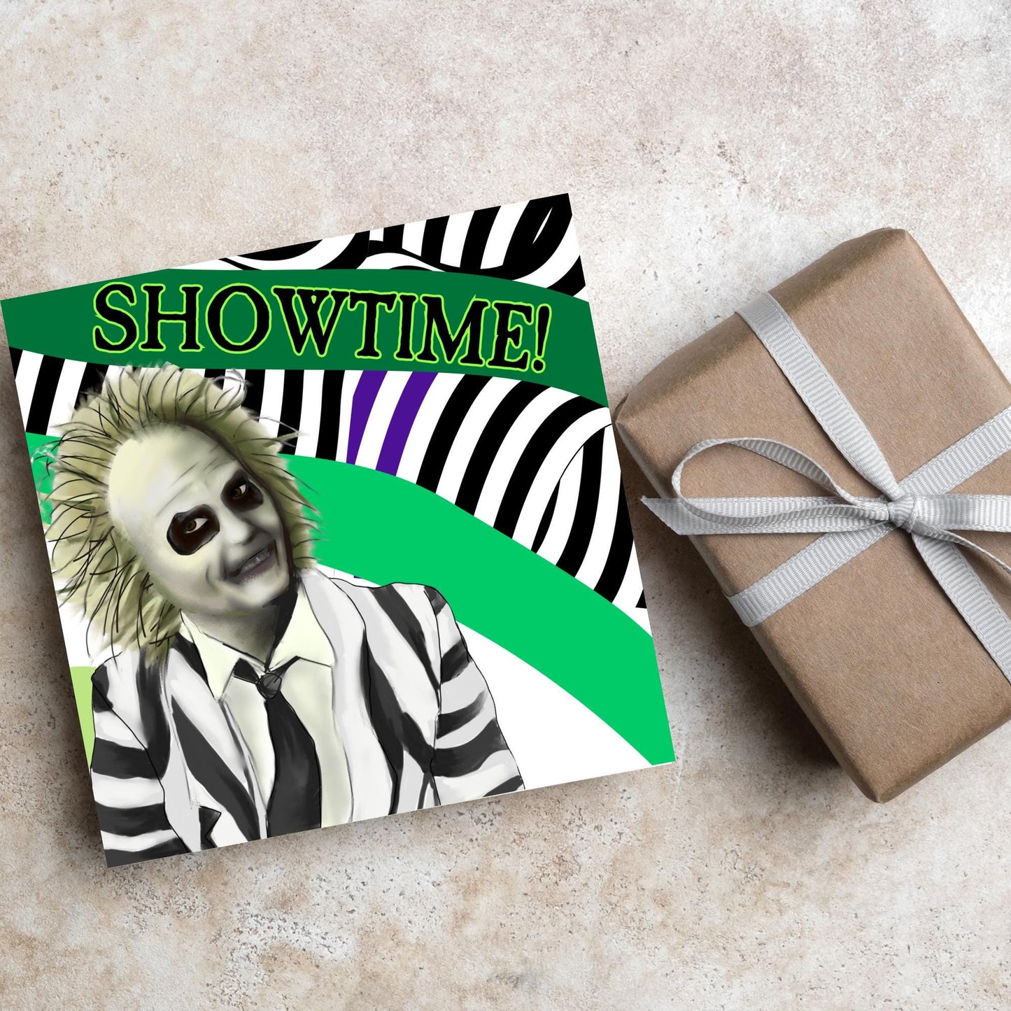 square greeting card with a drawn image of beetlejuice grinning in from of a black, green and purple striped background. On one of the biggest stripes is the word showtime in bold capital letters in a font similar to the movie title