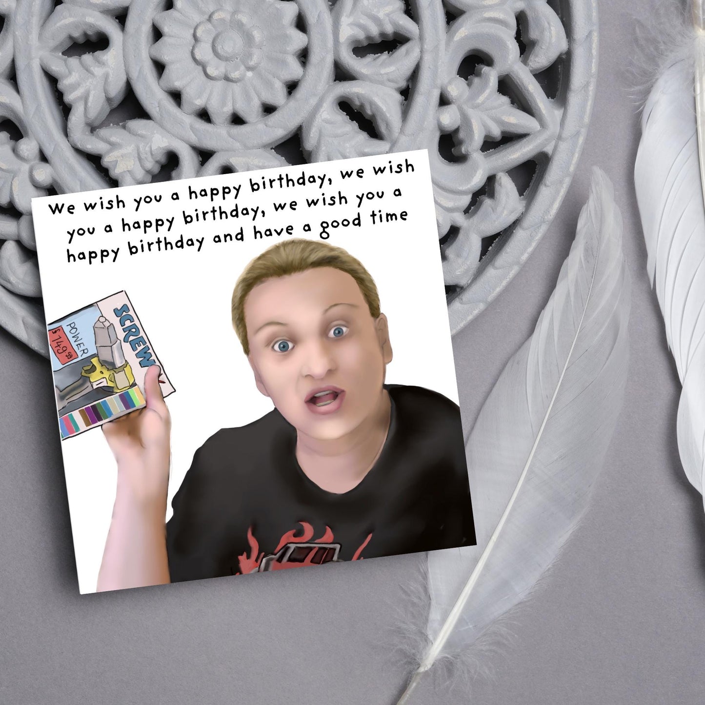 Square birthday card with a hand drawn image of Kerry from this country looking shocked and holding up a screwfix catalogue with the text we wish you a happy birthday, we wish you a happy birthday, we wish you a happy birthday and have a good time