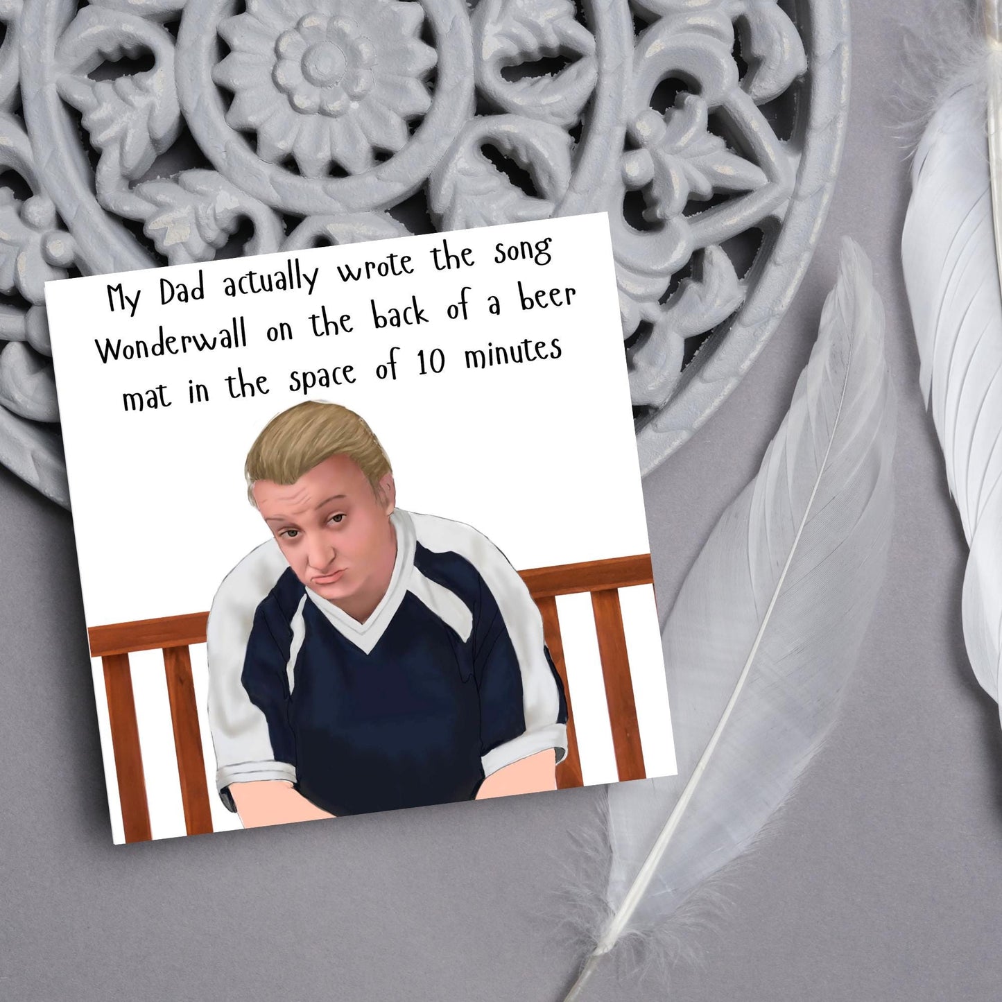 square card with a hand drawn image of kerry from this country sitting on a bench with handwritten style text above - My Dad actually wrote the song Wonderwall on the back of a beer mat in the space of 10 minutes