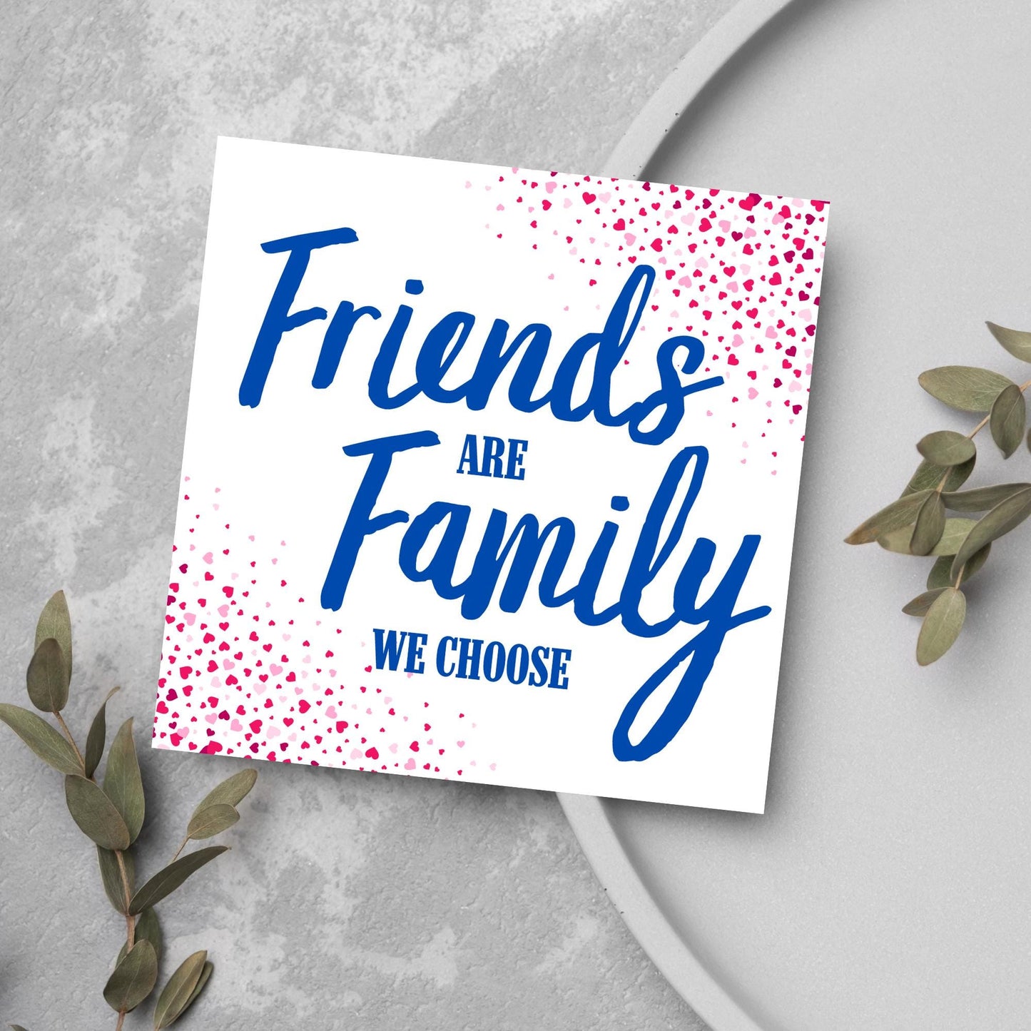 Square card with small hearts on opposite corners in shades of pink and blue writing in the centre that reads Friends are Family we choose