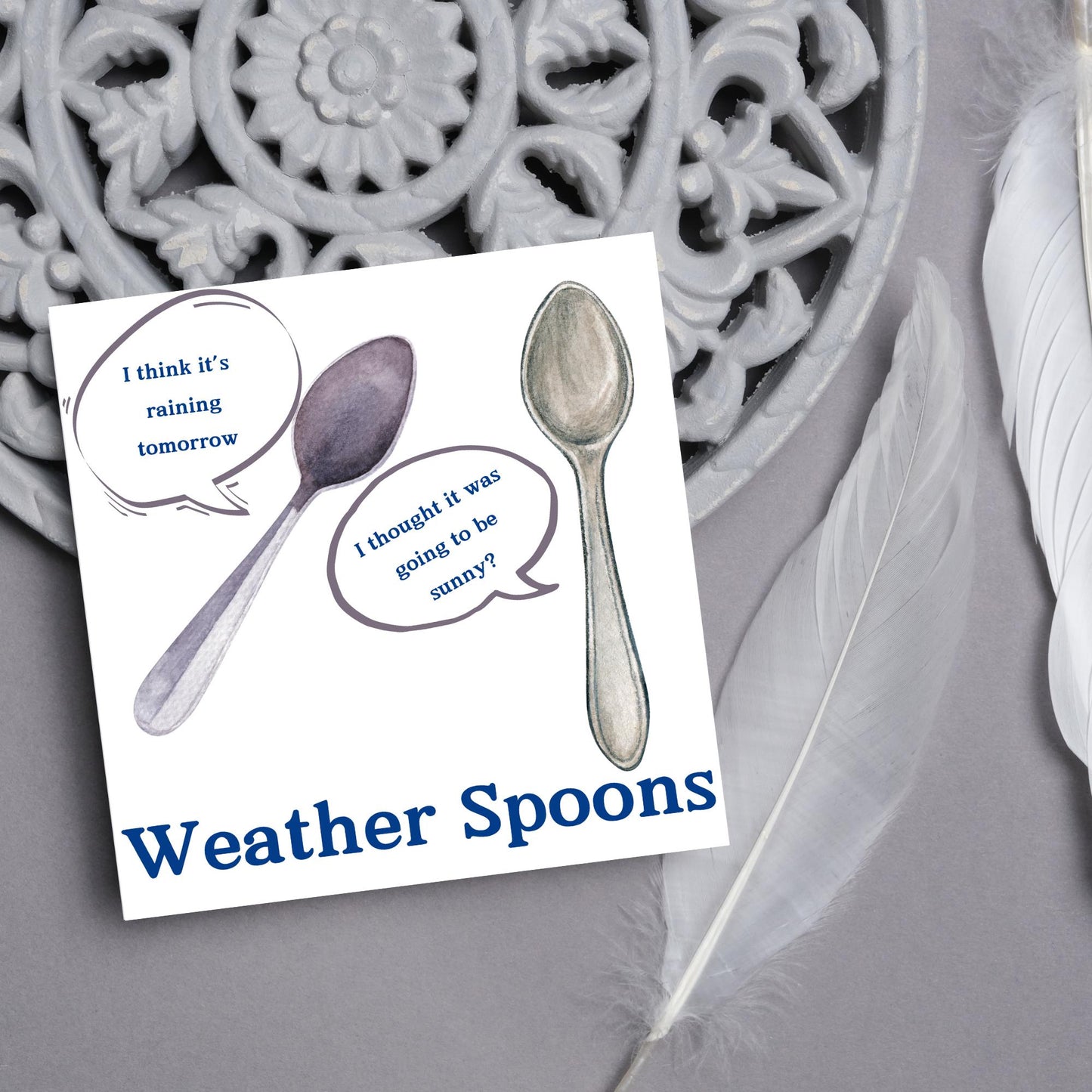 Image of a square greeting card. The card has a white backdrop with an illustration of two spoons with speech bubbles. One says I think it's raining tomorrow. The other says I thought it was going to be sunny? Underneath the illustration in a blue font is the text Weather Spoons