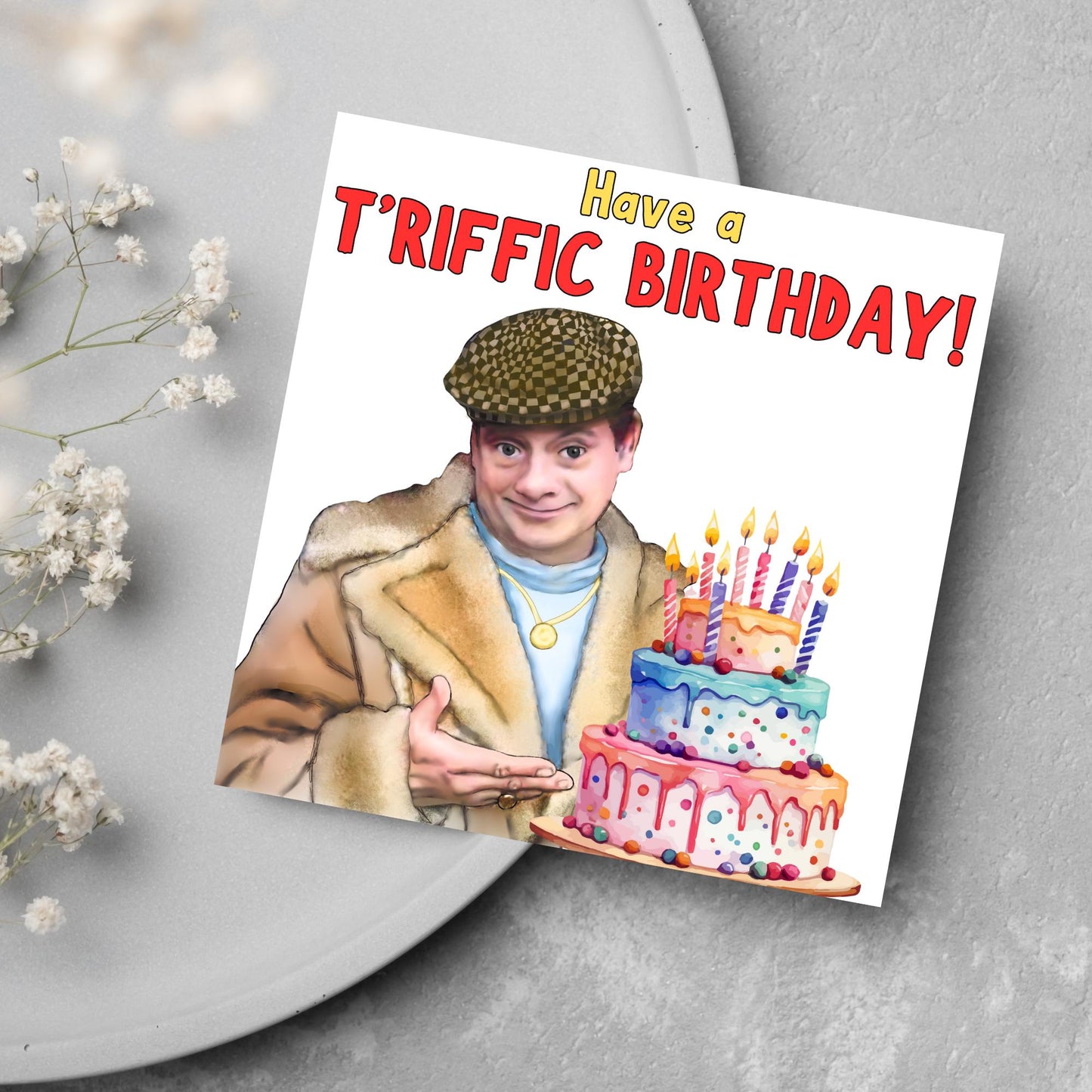 This square shaped card has an illustration of Del Boy from the British Sitcom Only Fools and Horses. The Card has Del Boy standing and pointing at an illustration of a triple tower birthday cake. The Caption reads have a T'riffic Birthday! The Caption is in a mustard yellow and ketchup red