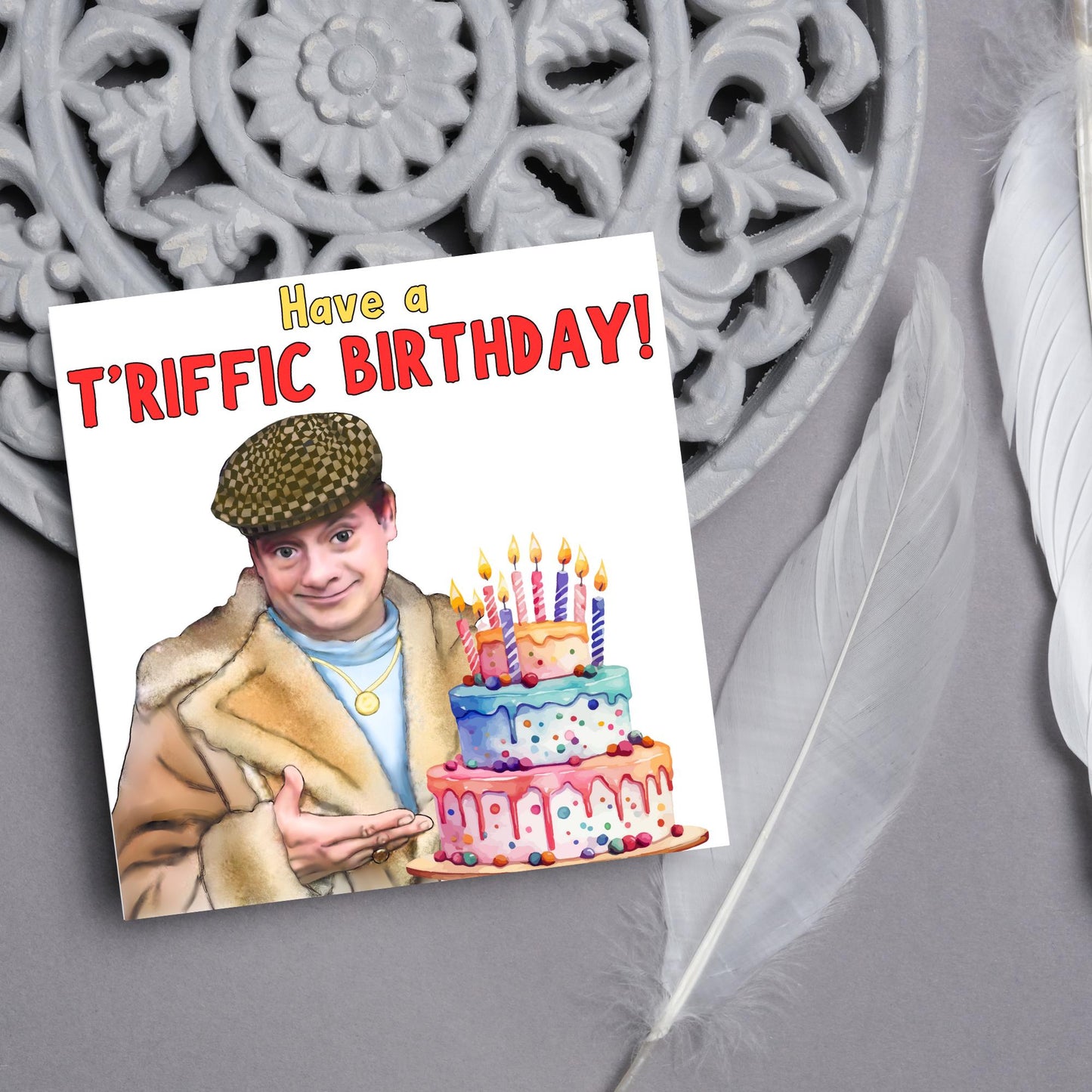 This square shaped card has an illustration of Del Boy from the British Sitcom Only Fools and Horses. The Card has Del Boy standing and pointing at an illustration of a triple tower birthday cake. The Caption reads have a T'riffic Birthday! The Caption is in a mustard yellow and ketchup red