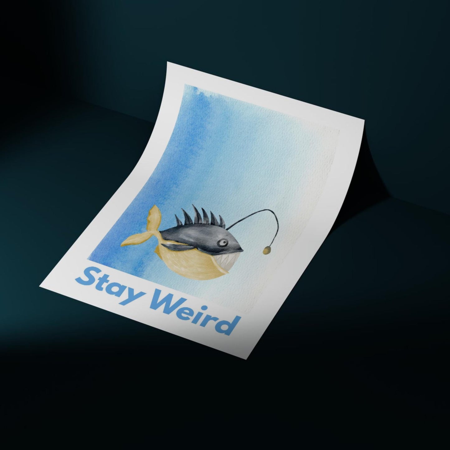 An A4 art print featuring a whimsical watercolor illustration of an anglerfish with a yellow tail and light lure, set against a blue gradient background. Below the fish, bold blue text reads "Stay Weird."