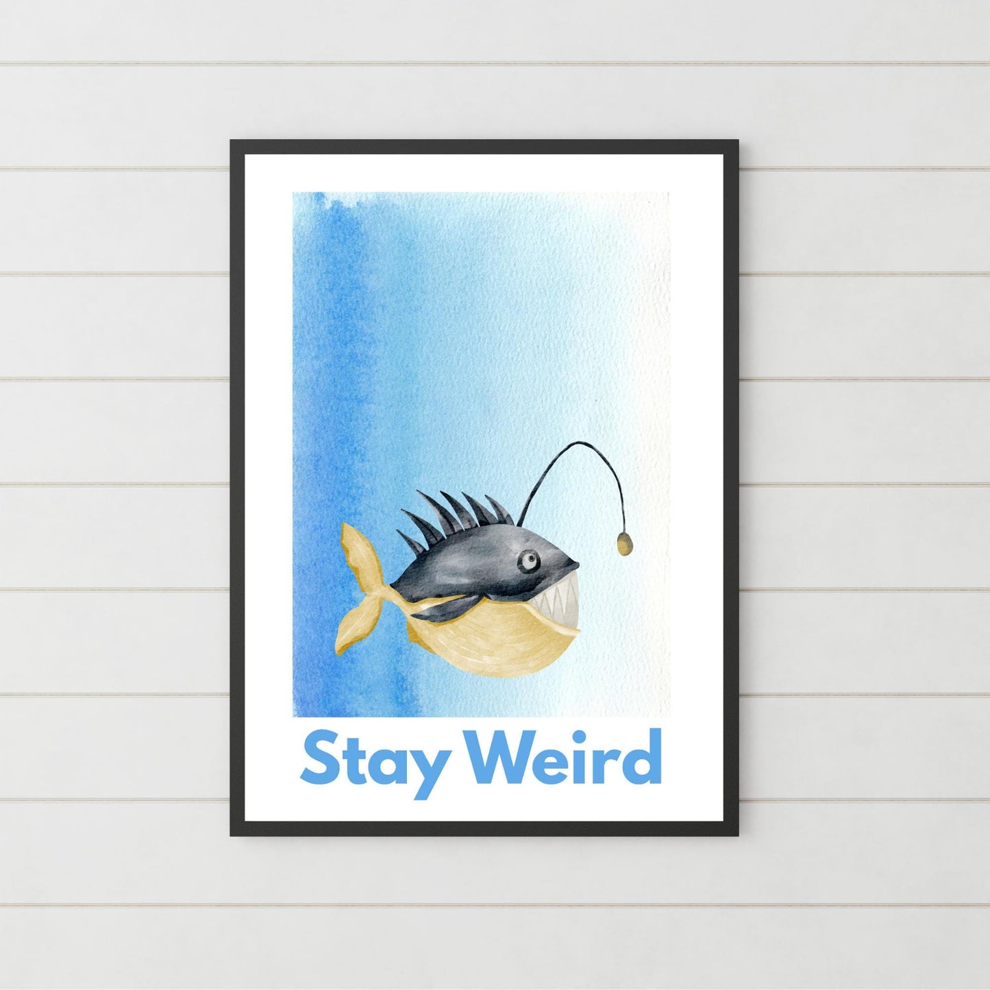 A framed A4 art print featuring a whimsical watercolor illustration of an anglerfish with a yellow tail and light lure, set against a blue gradient background. Below the fish, bold blue text reads "Stay Weird."