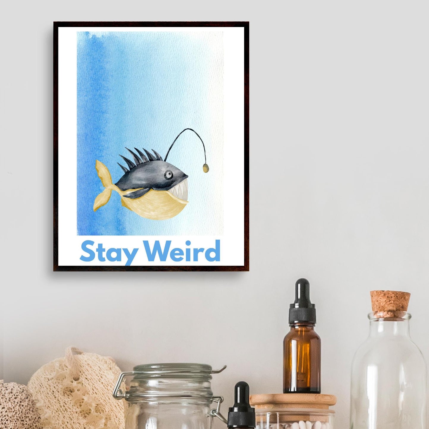 A framed A4 art print featuring a whimsical watercolor illustration of an anglerfish with a yellow tail and light lure, set against a blue gradient background. Below the fish, bold blue text reads "Stay Weird."