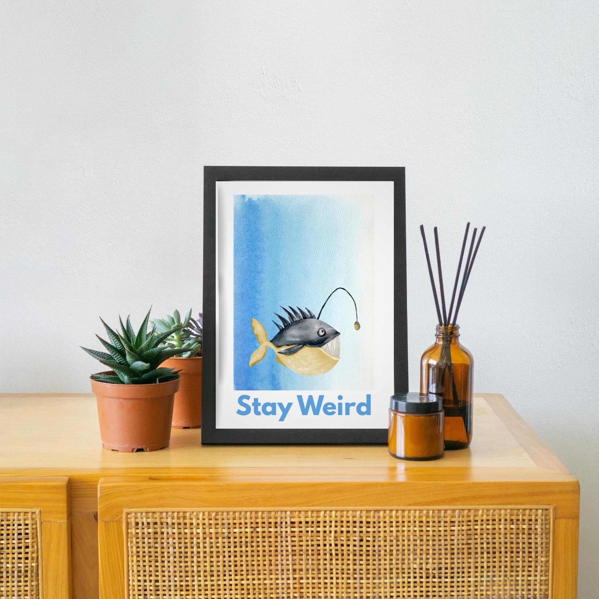 A framed A4 art print featuring a whimsical watercolor illustration of an anglerfish with a yellow tail and light lure, set against a blue gradient background. Below the fish, bold blue text reads "Stay Weird."
