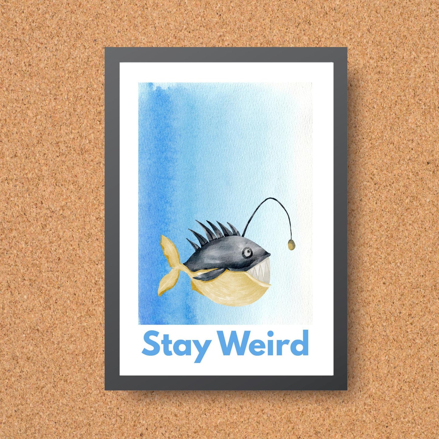 A framed A4 art print featuring a whimsical watercolor illustration of an anglerfish with a yellow tail and light lure, set against a blue gradient background. Below the fish, bold blue text reads "Stay Weird."