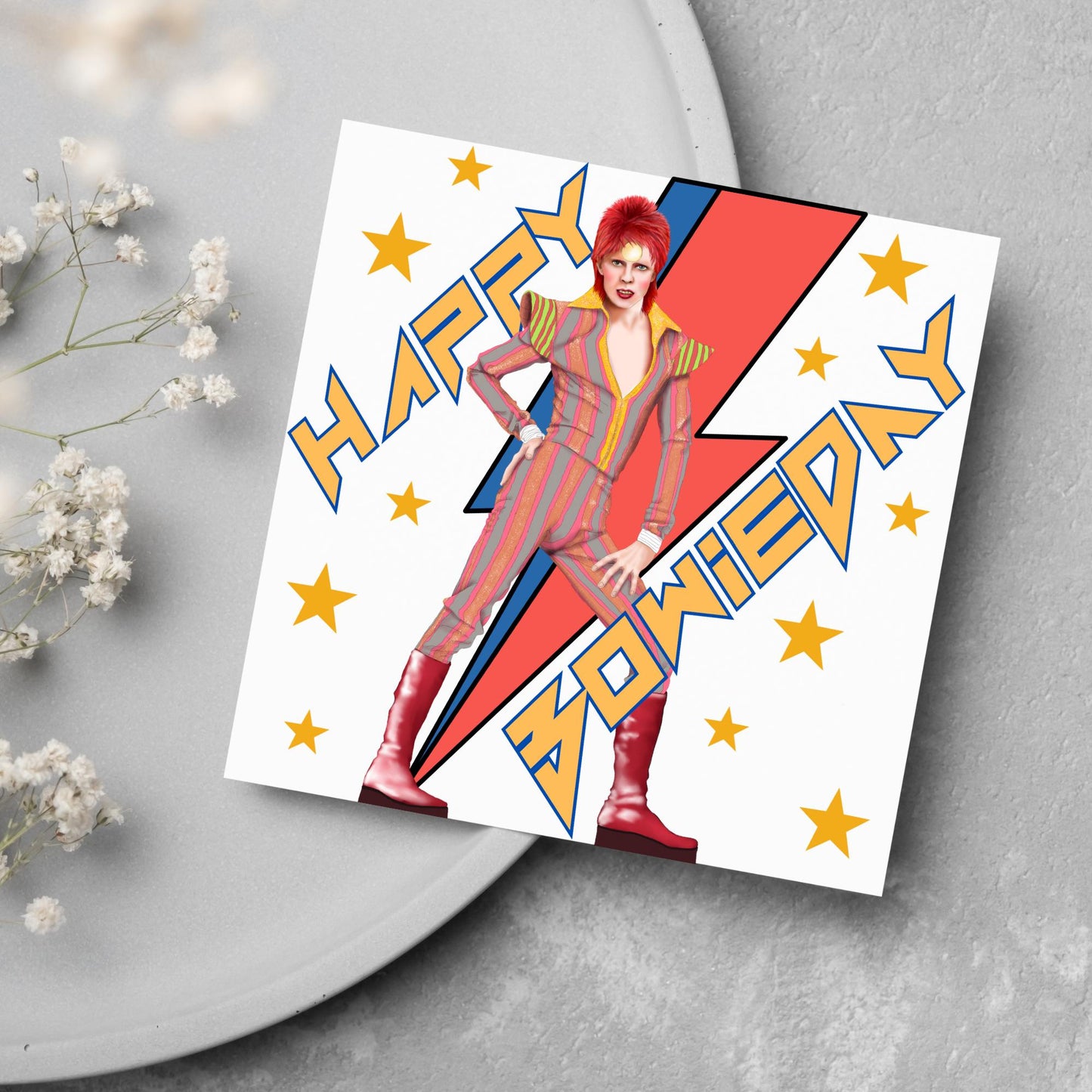 image of a square birthday card. Card has a white background with a vibrant illustration of David bowie with red hair and wearing a striped jumpsuit, behind him is the iconic Bowie lightning bolt in bright red and blue. Diagonal text reads Happy Bowieday in zigzag design bold yellow font. There are yellow stars dotted around the white background.