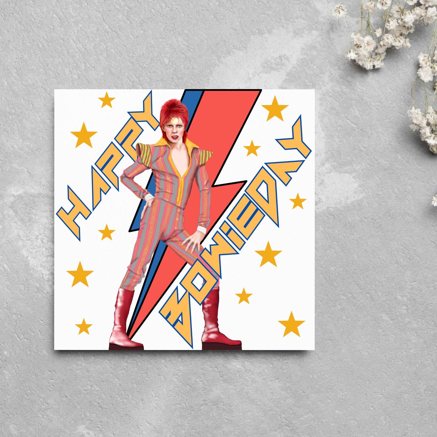 image of a square birthday card. Card has a white background with a vibrant illustration of David bowie with red hair and wearing a striped jumpsuit, behind him is the iconic Bowie lightning bolt in bright red and blue. Diagonal text reads Happy Bowieday in zigzag design bold yellow font. There are yellow stars dotted around the white background.