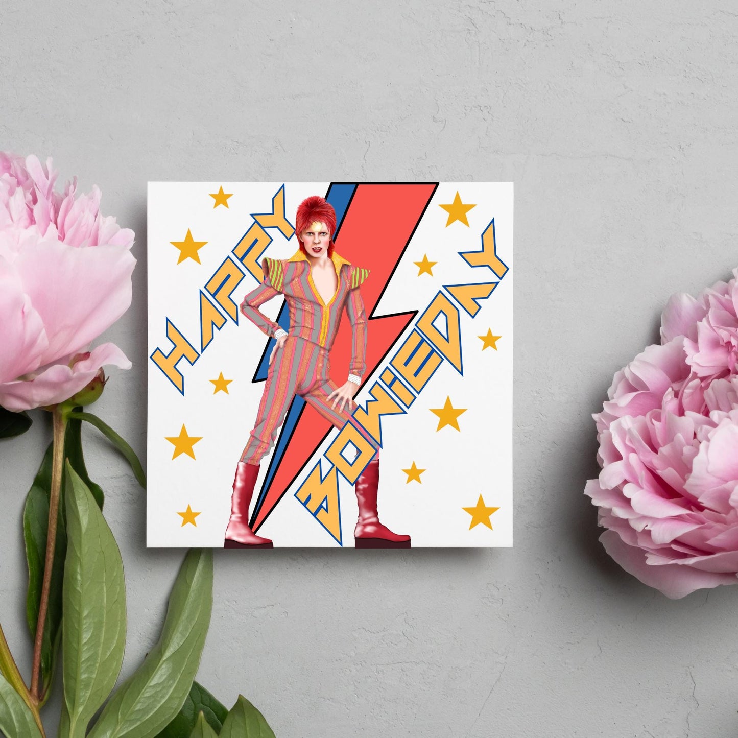 image of a square birthday card. Card has a white background with a vibrant illustration of David bowie with red hair and wearing a striped jumpsuit, behind him is the iconic Bowie lightning bolt in bright red and blue. Diagonal text reads Happy Bowieday in zigzag design bold yellow font. There are yellow stars dotted around the white background.