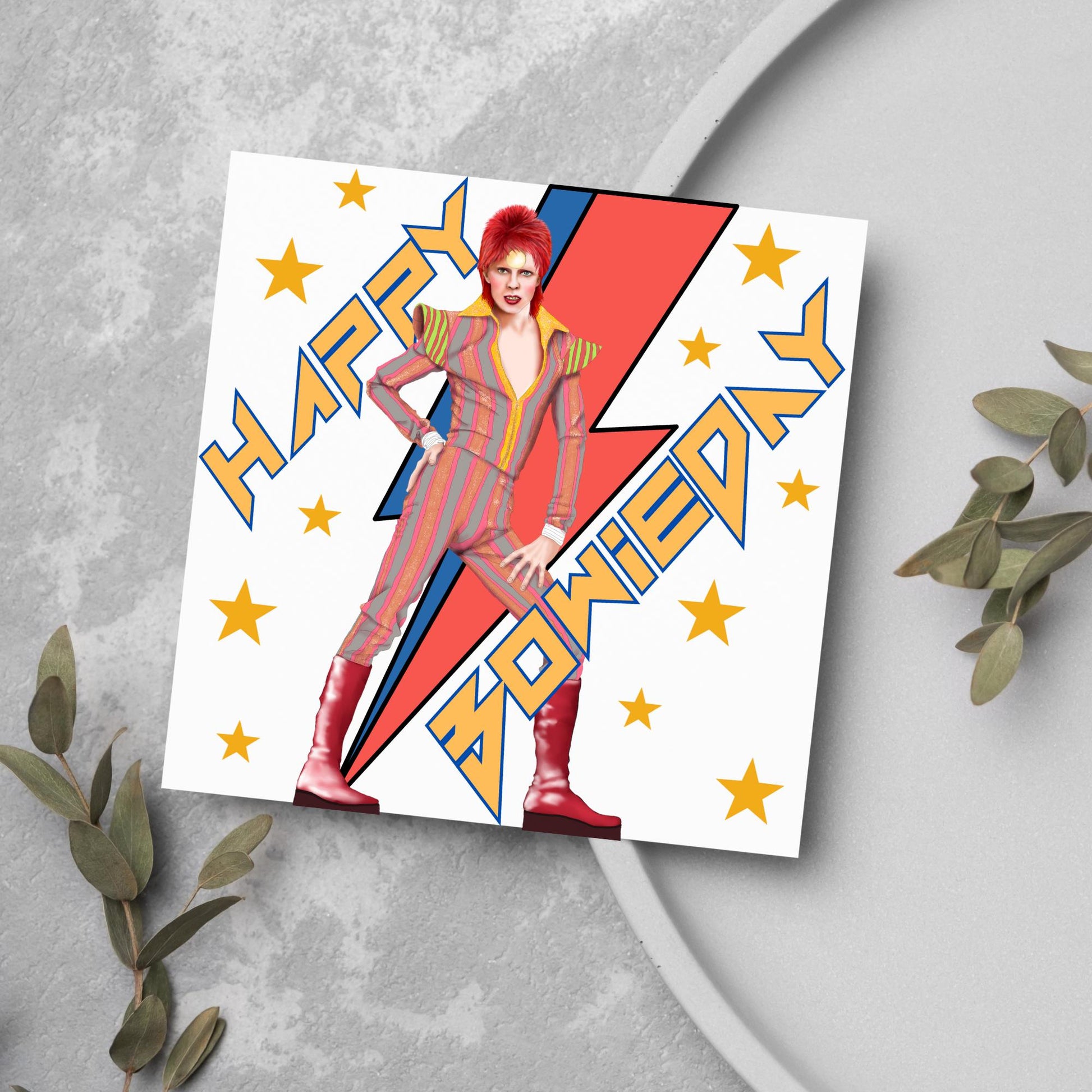 image of a square birthday card. Card has a white background with a vibrant illustration of David bowie with red hair and wearing a striped jumpsuit, behind him is the iconic Bowie lightning bolt in bright red and blue. Diagonal text reads Happy Bowieday in zigzag design bold yellow font. There are yellow stars dotted around the white background.