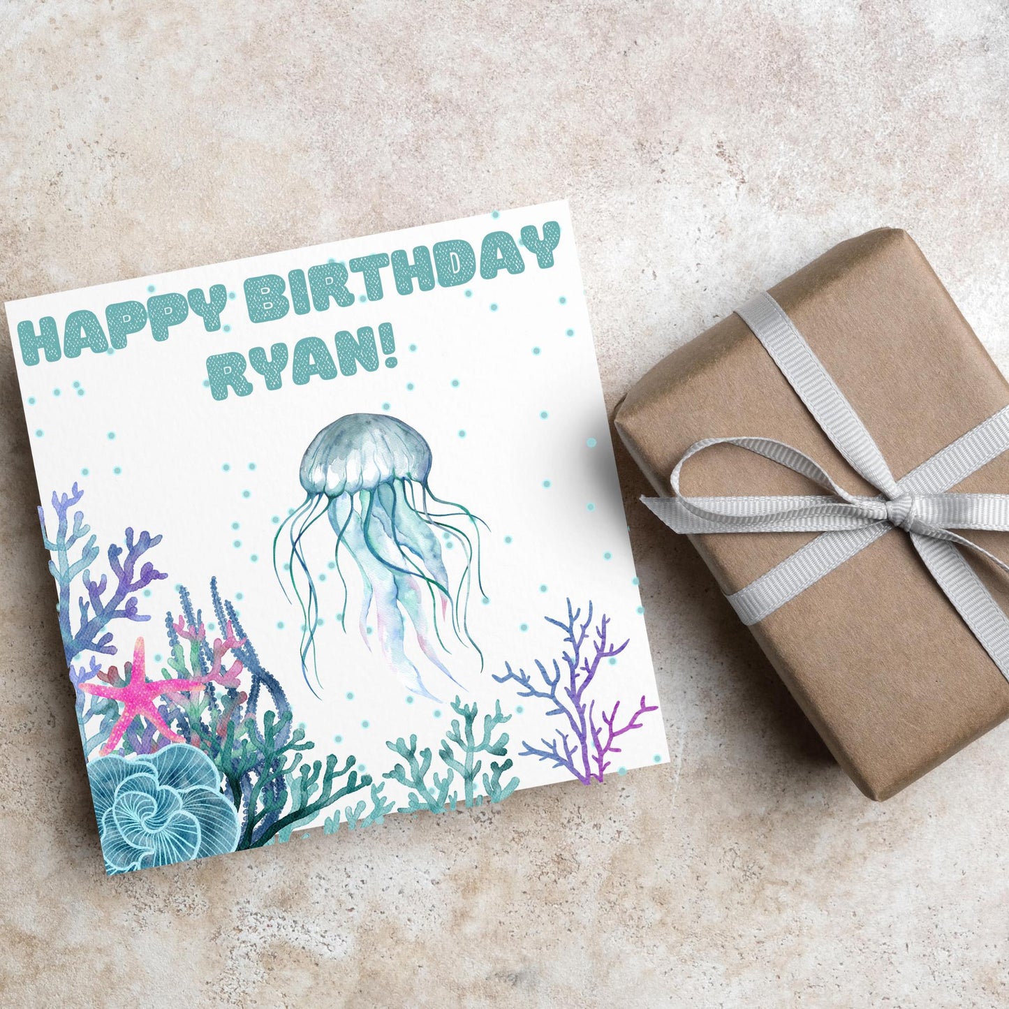 Image of a square Birthday card. Card has a white background and a watercolour style jellyfish amongst coral and seaweed. All the colours are blues, greens, pinks and purples. At the top of the card is bold blue text that says happy birthday ryan which can be personalised to any name.