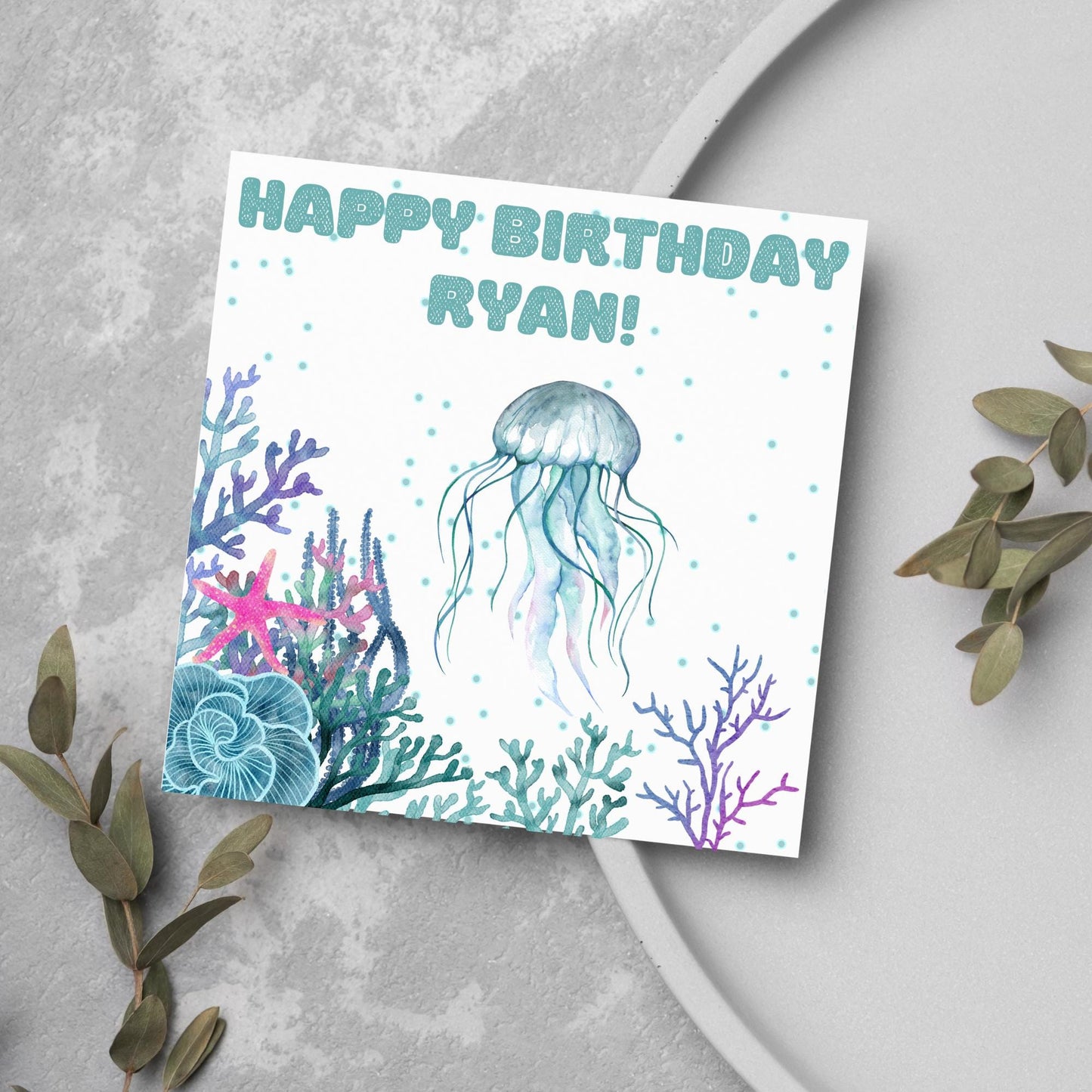 Image of a square Birthday card. Card has a white background and a watercolour style jellyfish amongst coral and seaweed. All the colours are blues, greens, pinks and purples. At the top of the card is bold blue text that says happy birthday ryan which can be personalised to any name.